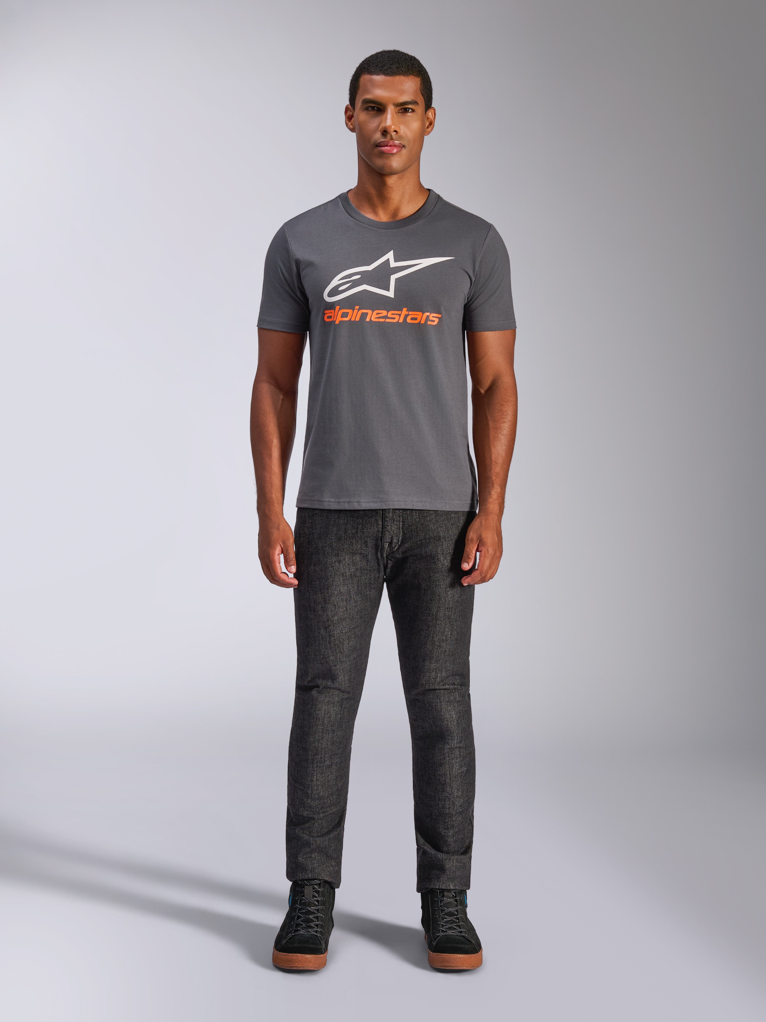 Alpinestars Always 2.0 CSF Tee, t-shirt de manga curta, cinzento carvão com logótipo branco e texto laranja, vestida por um modelo com calças de ganga escuras, streetwear casual de motorsport