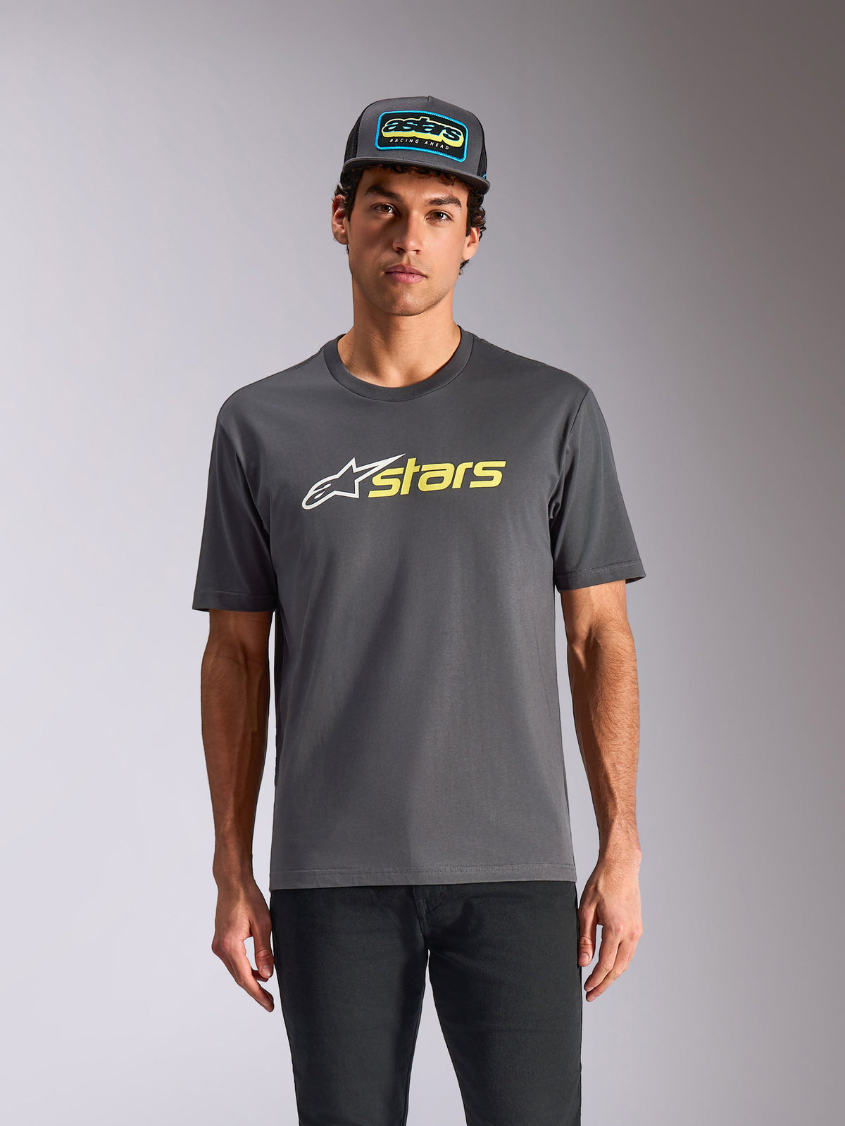 Alpinestars Blaze 2.0 CSF Tee, t-shirt de manga curta, cinzento carvão, logótipo de estrela branco e gráfico 'stars' em amarelo Hi-Vis, usada com boné trucker a condizer e calças pretas, streetwear casual de desportos motorizados