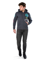 Alpinestars Variegate Hoodie, zip-front fleece hoodie, Gunmetal gray, worn by model with black jeans and sneakers, featuring teal 'ASTARS' branding and colorful logo art on sleeves and chest, casual motorsport-inspired streetwear