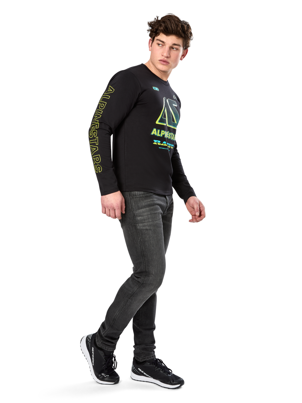 Homem a usar t-shirt de manga comprida Alpinestars Ayess CSF, preta, com letras amarelas na manga e gráficos vibrantes em lima e ciano no peito, combinada com calças de ganga cinzento-escuro e sapatilhas pretas, estilo streetwear casual inspirado no desporto motorizado.