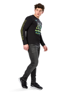 Homem a usar t-shirt de manga comprida Alpinestars Ayess CSF, preta, com letras amarelas na manga e gráficos vibrantes em lima e ciano no peito, combinada com calças de ganga cinzento-escuro e sapatilhas pretas, estilo streetwear casual inspirado no desporto motorizado.
