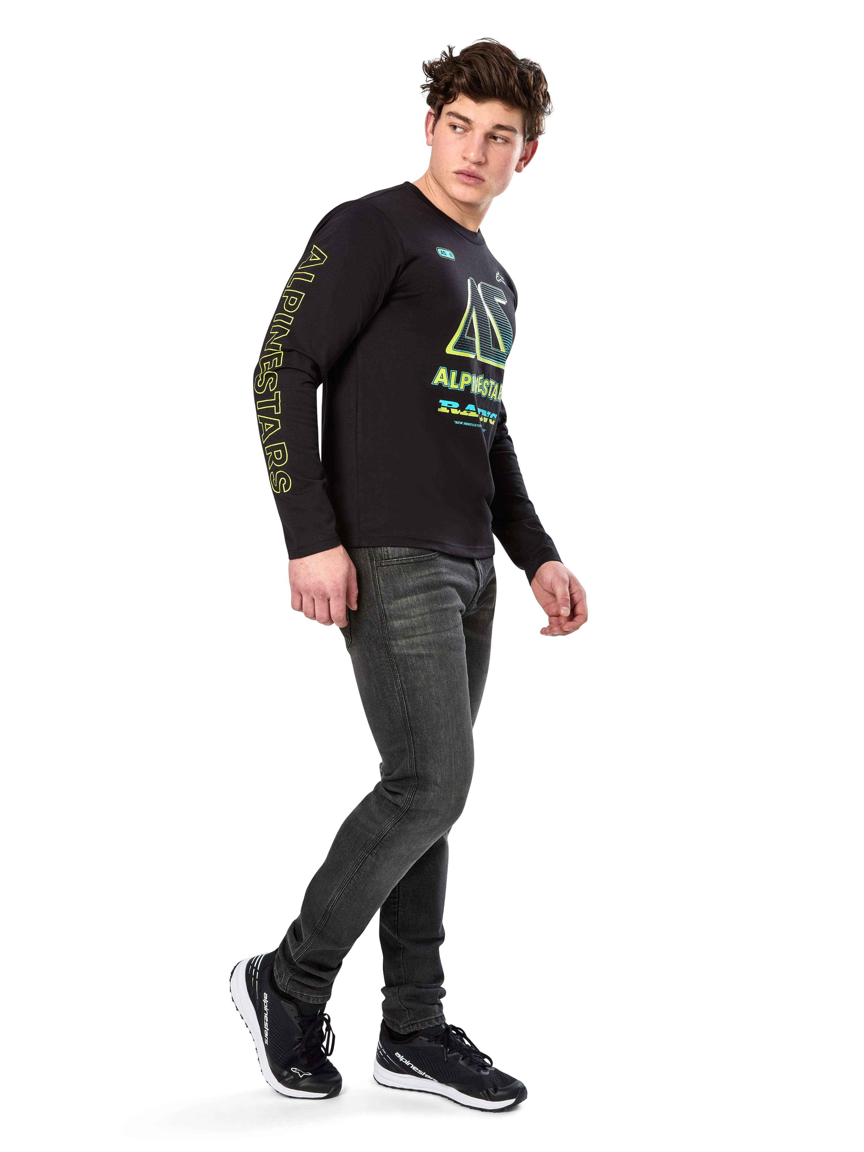 Homem a usar t-shirt de manga comprida Alpinestars Ayess CSF, preta, com letras amarelas na manga e gráficos vibrantes em lima e ciano no peito, combinada com calças de ganga cinzento-escuro e sapatilhas pretas, estilo streetwear casual inspirado no desporto motorizado.
