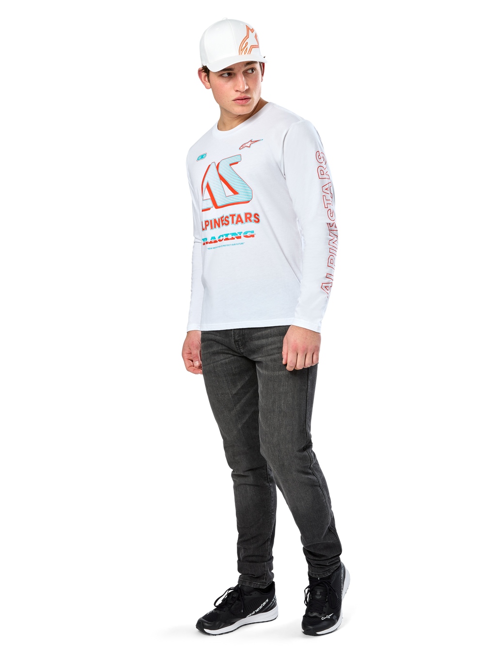Alpinestars Ayess CSF Tee, T-shirt de manga comprida branca, com grandes grafismos de corrida CSF em azul e vermelho no peito e logótipo Alpinestars vermelho na manga, usada com calças de ganga escuras e um boné Alpinestars branco, streetwear casual de desportos motorizados
