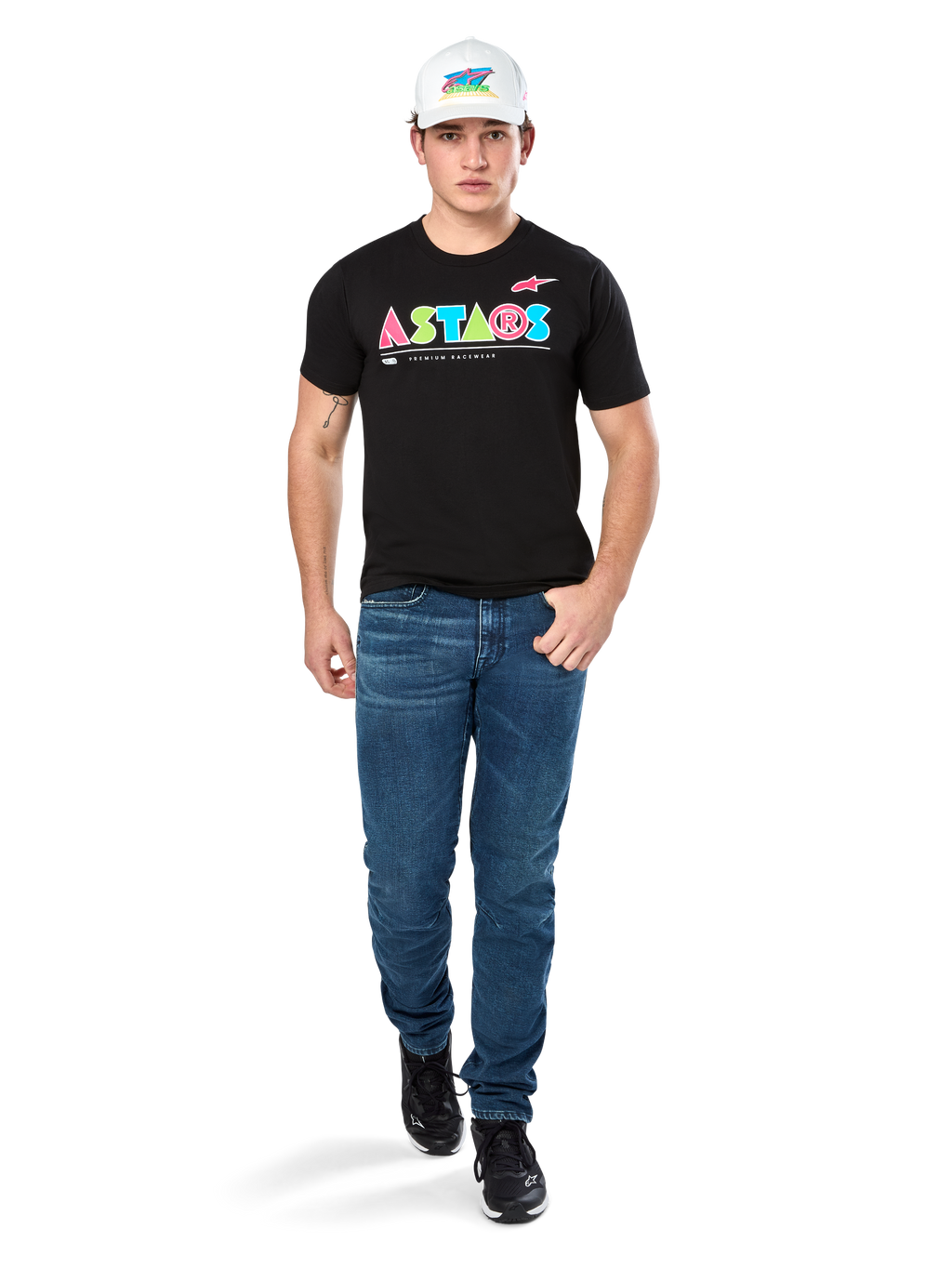 Modelo masculino a usar a T-shirt Alpinestars Cereal CSF, t-shirt preta de manga curta com logótipo ASTARS geométrico e colorido, combinada com calças de ganga azuis e boné branco, streetwear casual inspirado no desporto motorizado.