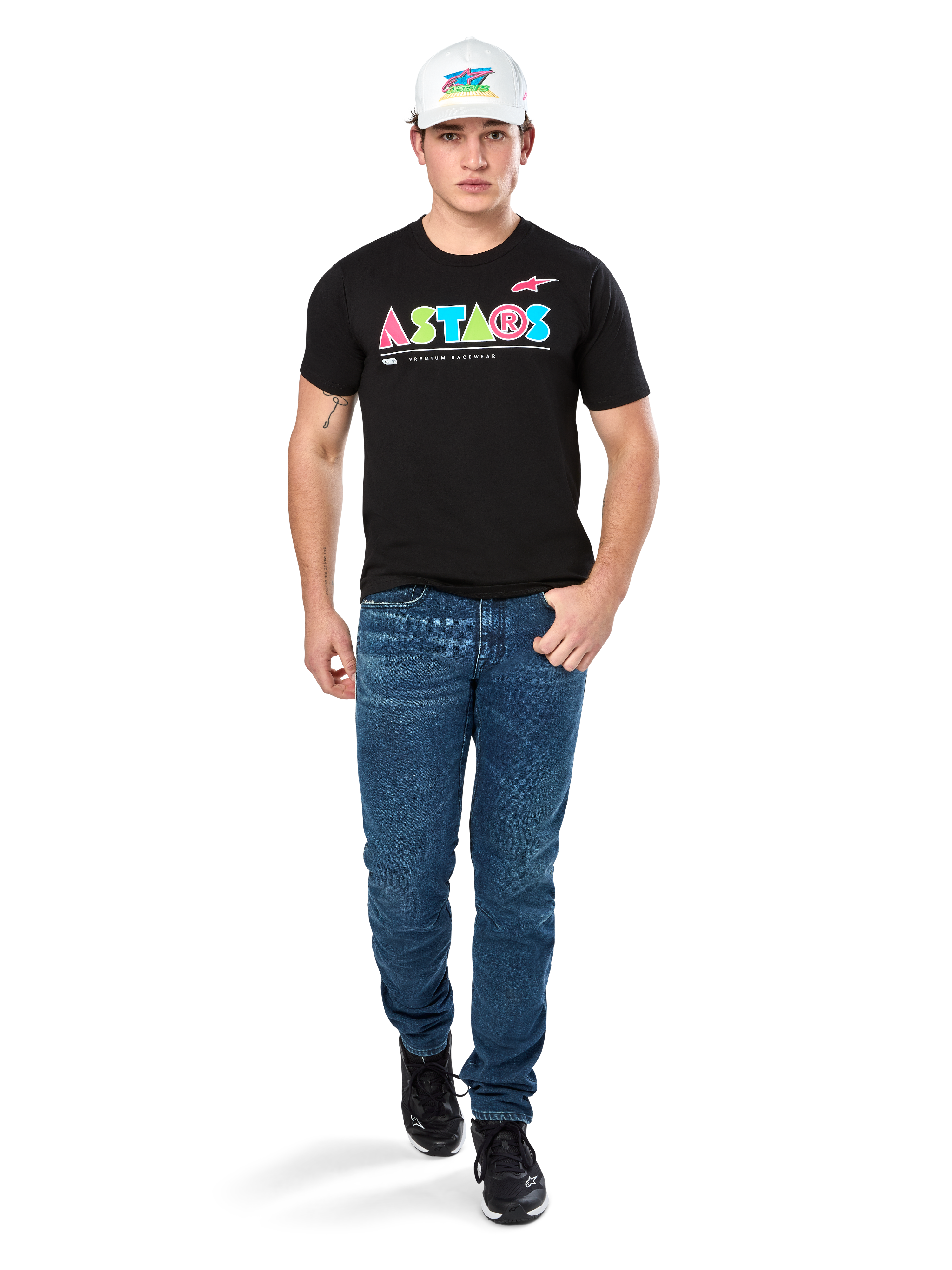 Modelo masculino a usar a T-shirt Alpinestars Cereal CSF, t-shirt preta de manga curta com logótipo ASTARS geométrico e colorido, combinada com calças de ganga azuis e boné branco, streetwear casual inspirado no desporto motorizado.