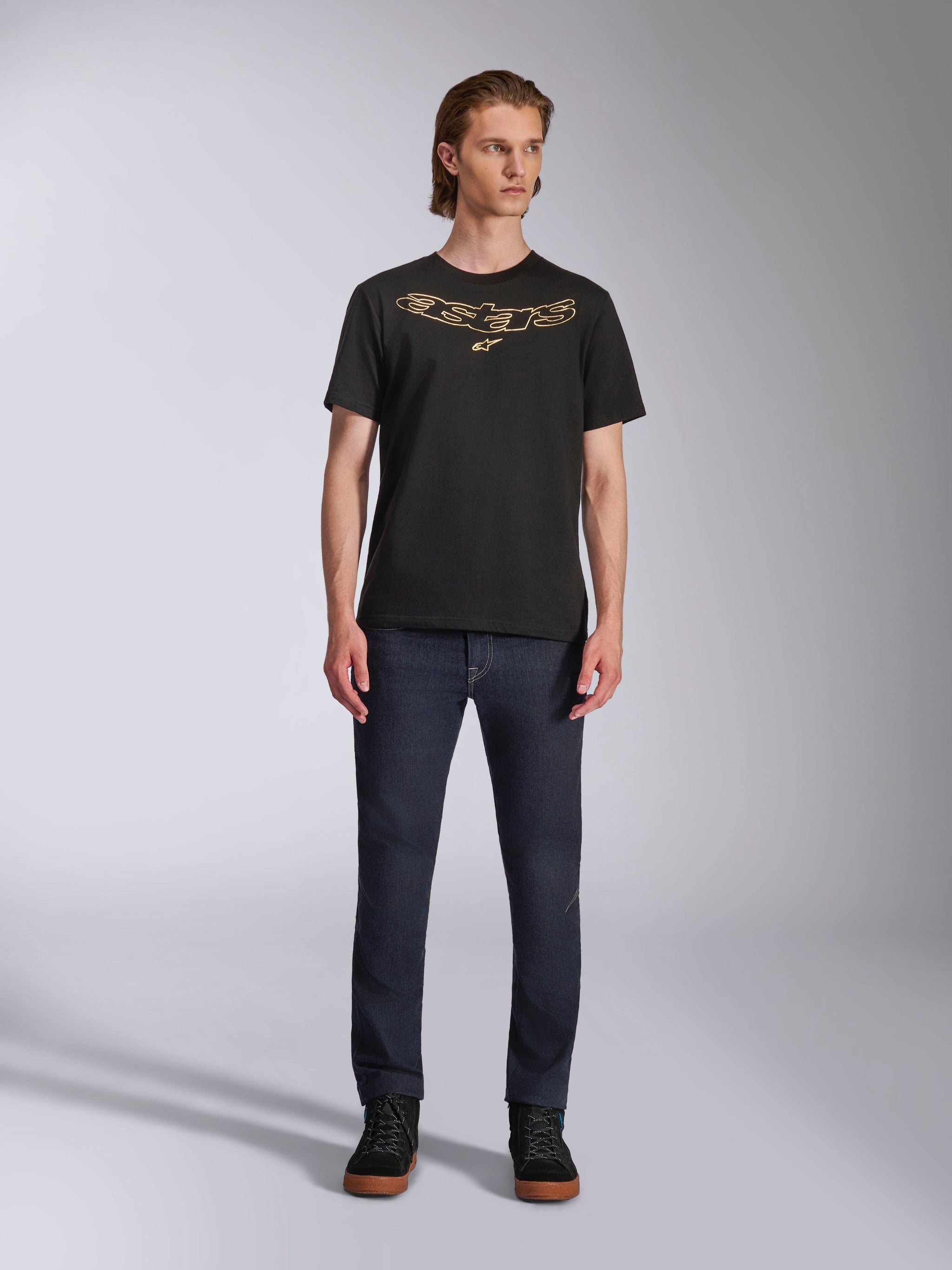 Alpinestars Luxo CSF Tee, t-shirt de manga curta, preta, logótipo Astars no peito com contorno bege, usada por modelo com calças de ganga escuras e sapatilhas pretas de cano alto, streetwear casual de desporto motorizado