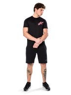 Modelo a usar a t-shirt Alpinestars Attrition Performance, t-shirt de performance de manga curta, preta com logótipo Astars cor-de-rosa no peito, combinada com calções pretos e ténis, roupa desportiva casual inspirada no desporto motorizado
