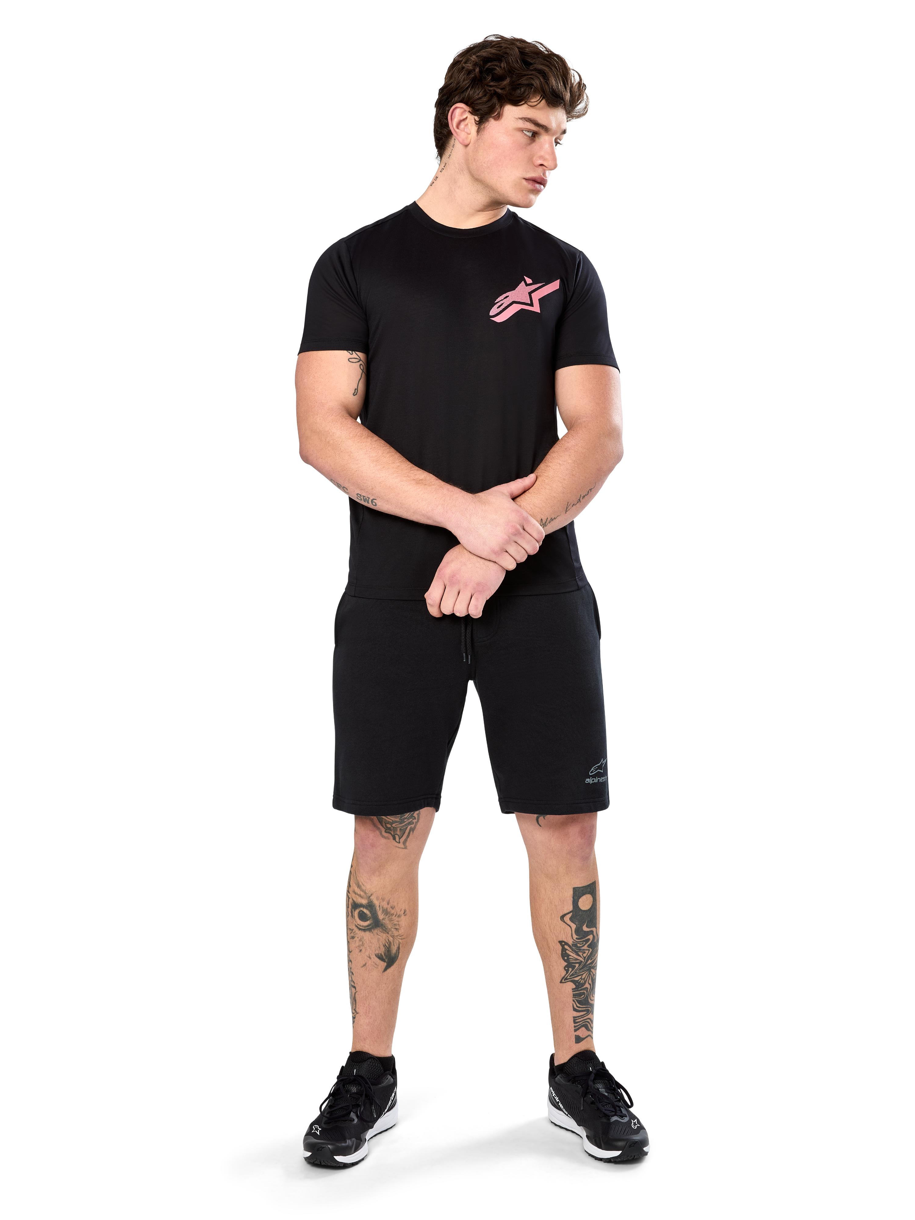Modelo a usar a t-shirt Alpinestars Attrition Performance, t-shirt de performance de manga curta, preta com logótipo Astars cor-de-rosa no peito, combinada com calções pretos e ténis, roupa desportiva casual inspirada no desporto motorizado