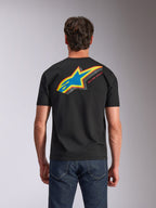 T-shirt Alpinestars Angler CSF, de manga curta, preta, vista de trás mostrando o logótipo Astars vibrante e multicolor com preenchimento perfurado e o texto 'RACING AHEAD', usada com calças de ganga azuis, vestuário casual inspirado no desporto motorizado