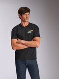 Alpinestars Angler CSF Tee, T-shirt de manga curta, preta, com um logótipo Astars vibrante e multicolor no peito, usada por um modelo com os braços cruzados e calças de ganga azuis, streetwear casual inspirado no desporto motorizado