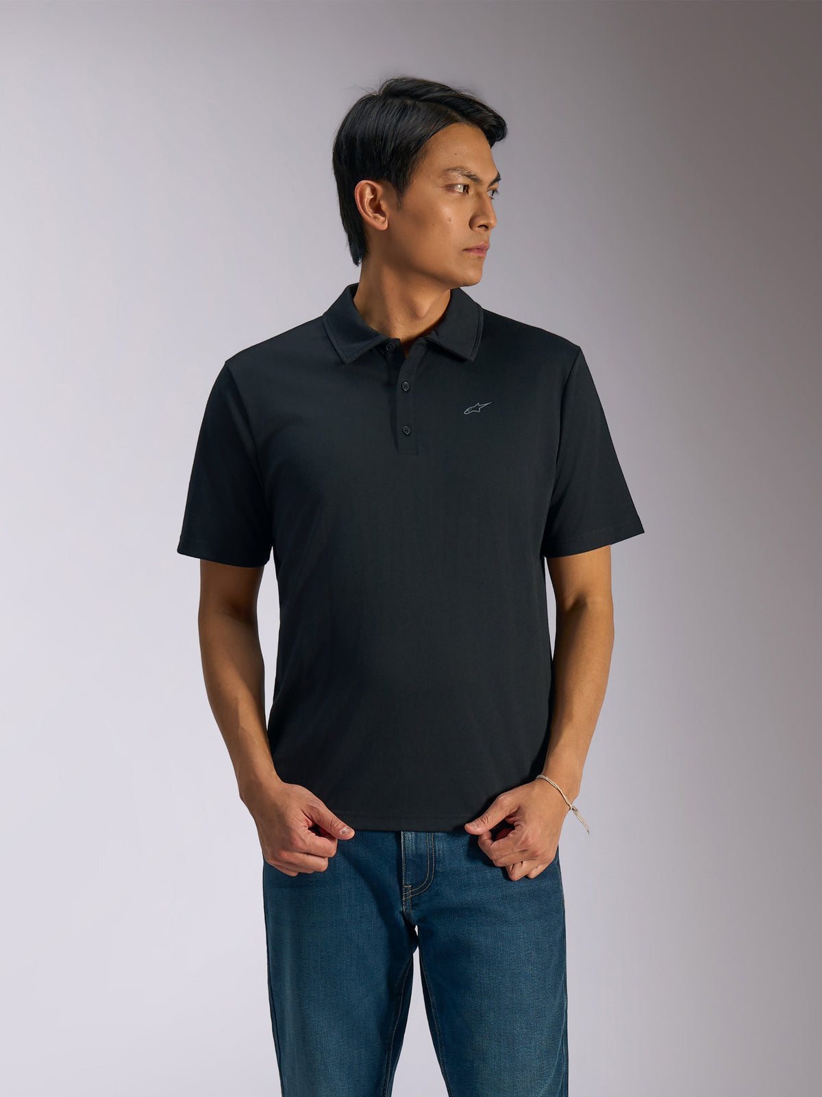 Alpinestars Impetus Sport Polo, polo de manga curta preto, carcela de três botões e gola própria, pequeno logótipo Astars cinzento no peito esquerdo, usado com calças de ganga azuis, vestuário desportivo casual de inspiração motorsport