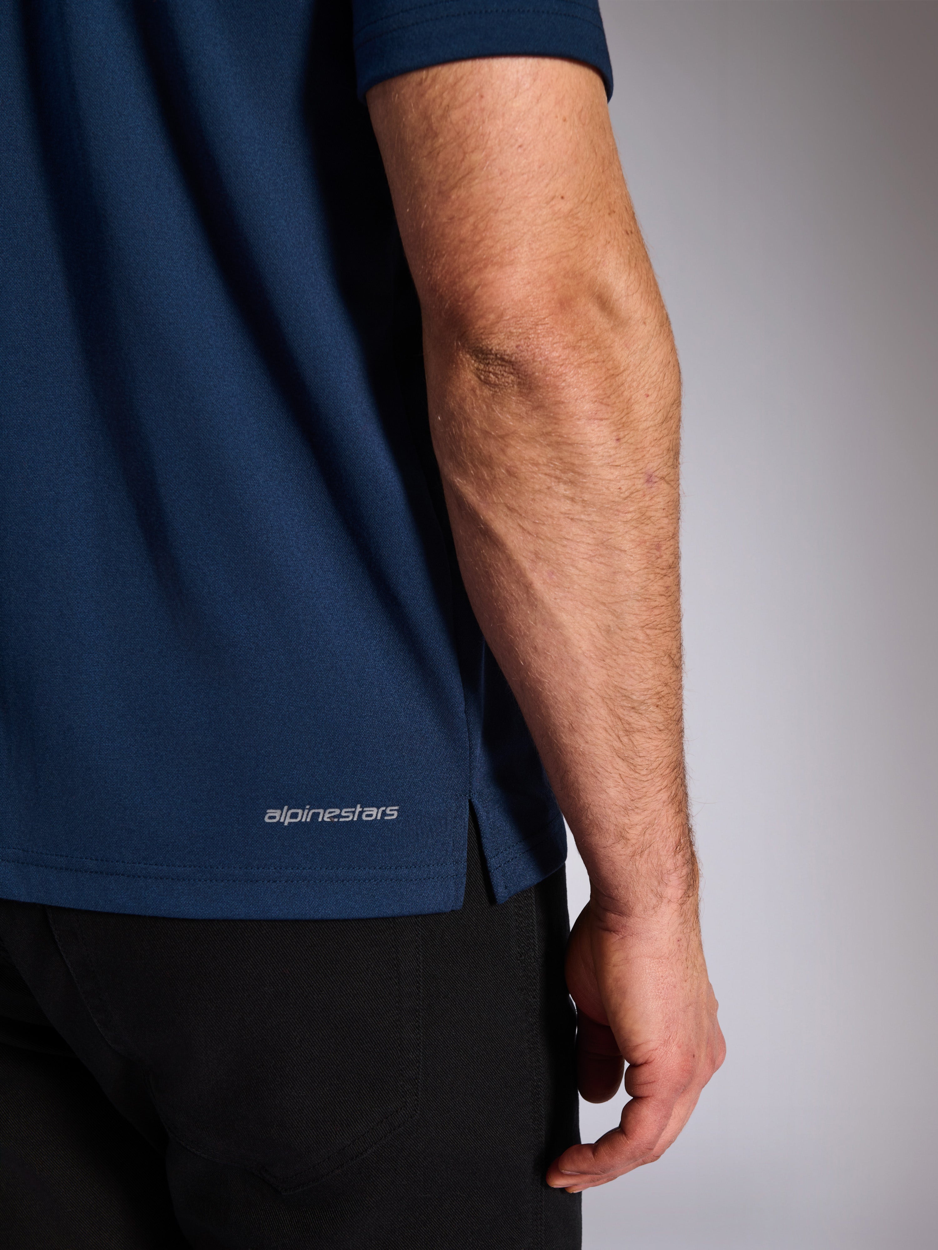 Alpinestars Impetus Sport Polo, azul marinho, grande plano da bainha inferior mostrando o logótipo Alpinestars e a abertura lateral, textura de malha mock-mesh, roupa desportiva casual de motociclismo