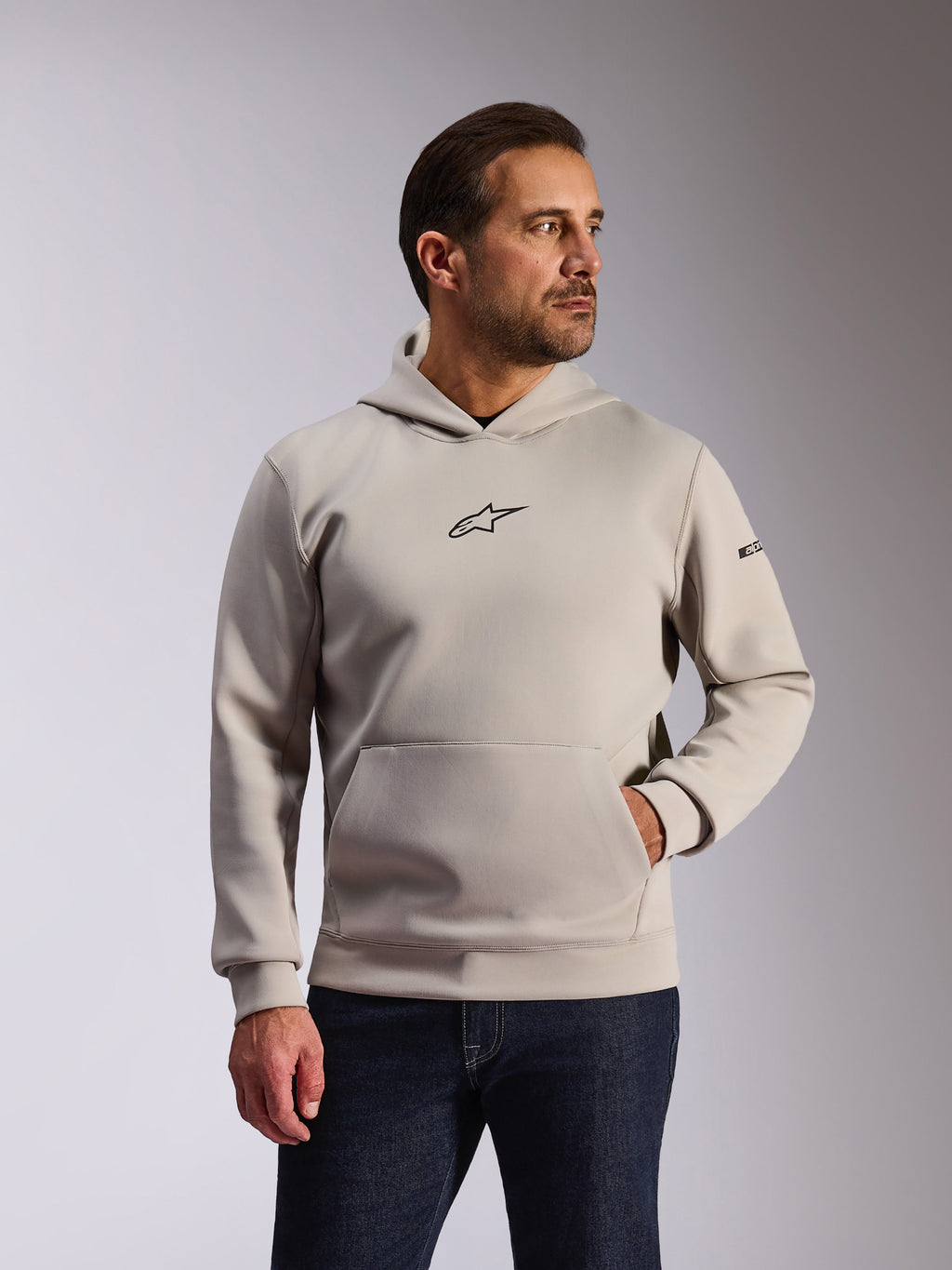 Alpinestars Insight Hoodie, sweatshirt casual, cinzento cimento, design de vestir pela cabeça com bolso canguru e pequeno logótipo Astars preto no peito, mangas compridas com logótipo Alpinestars no braço esquerdo, vestido por um modelo com calças de ganga escuras, vestuário de desporto motorizado lifestyle