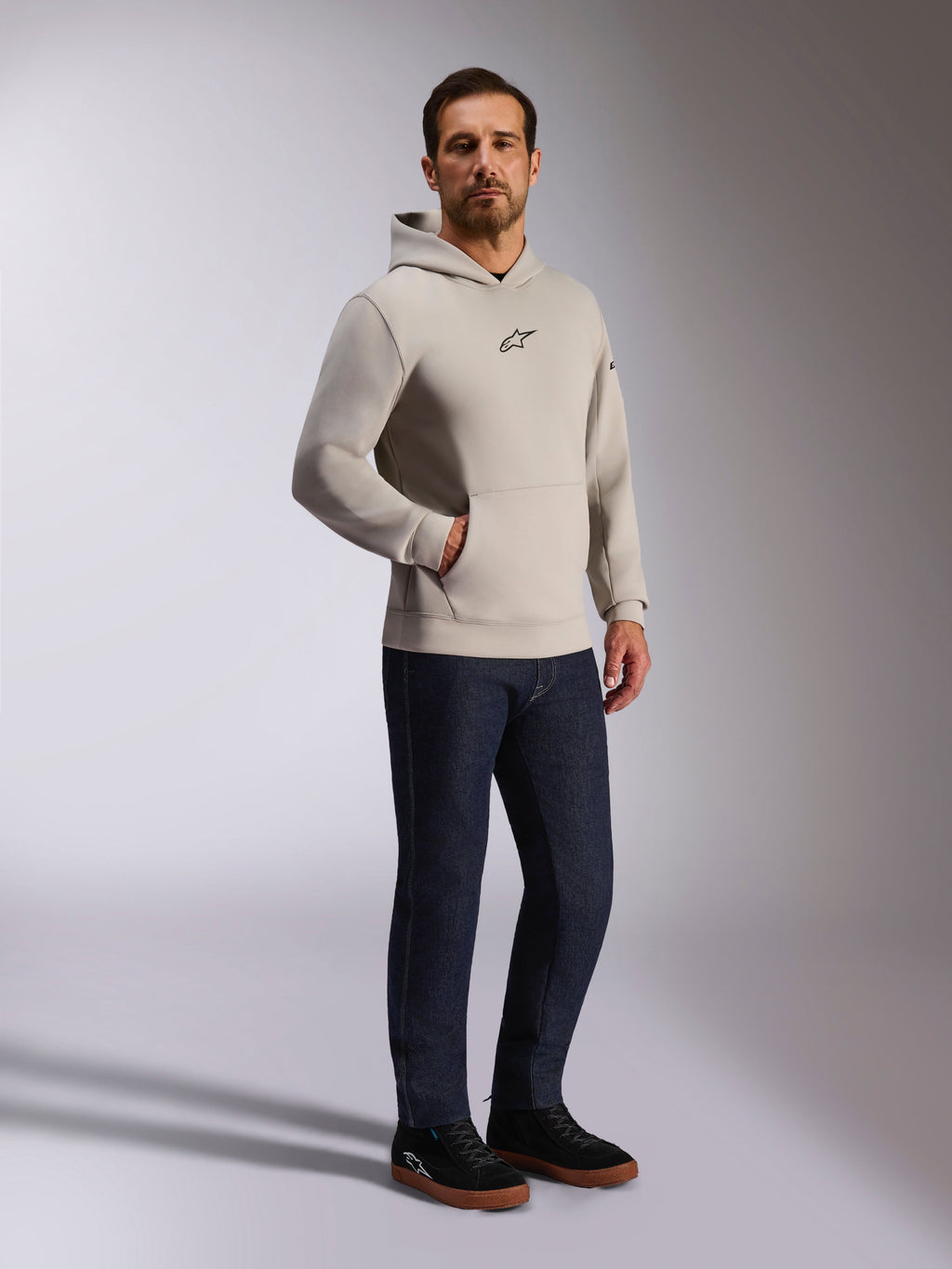 Alpinestars Insight Hoodie, casual streetwear, cinzento Cement com logótipo Astars preto no peito, design pullover com bolso canguru, usado com calças de ganga azul escuro e ténis pretos, estilo moderno inspirado no desporto motorizado