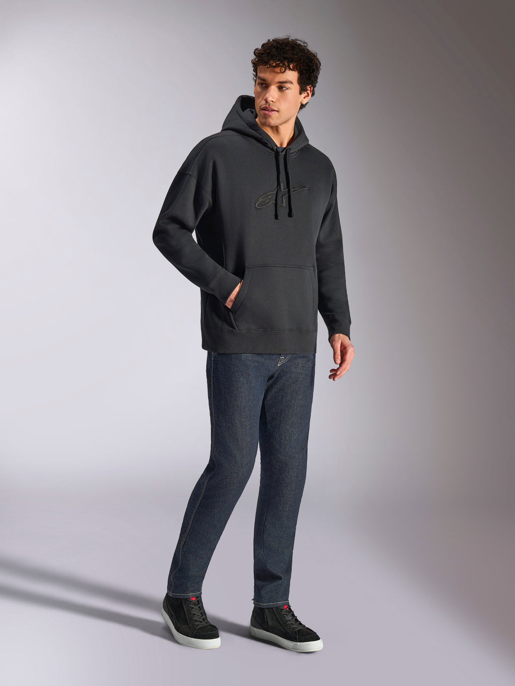 Alpinestars Emergent Oversized Hoodie, black casual streetwear, tonal 3D embroidered Astars logo on chest, kangaroo pocket, oversized fit, worn by male model with dark blue denim jeans and black sneakers