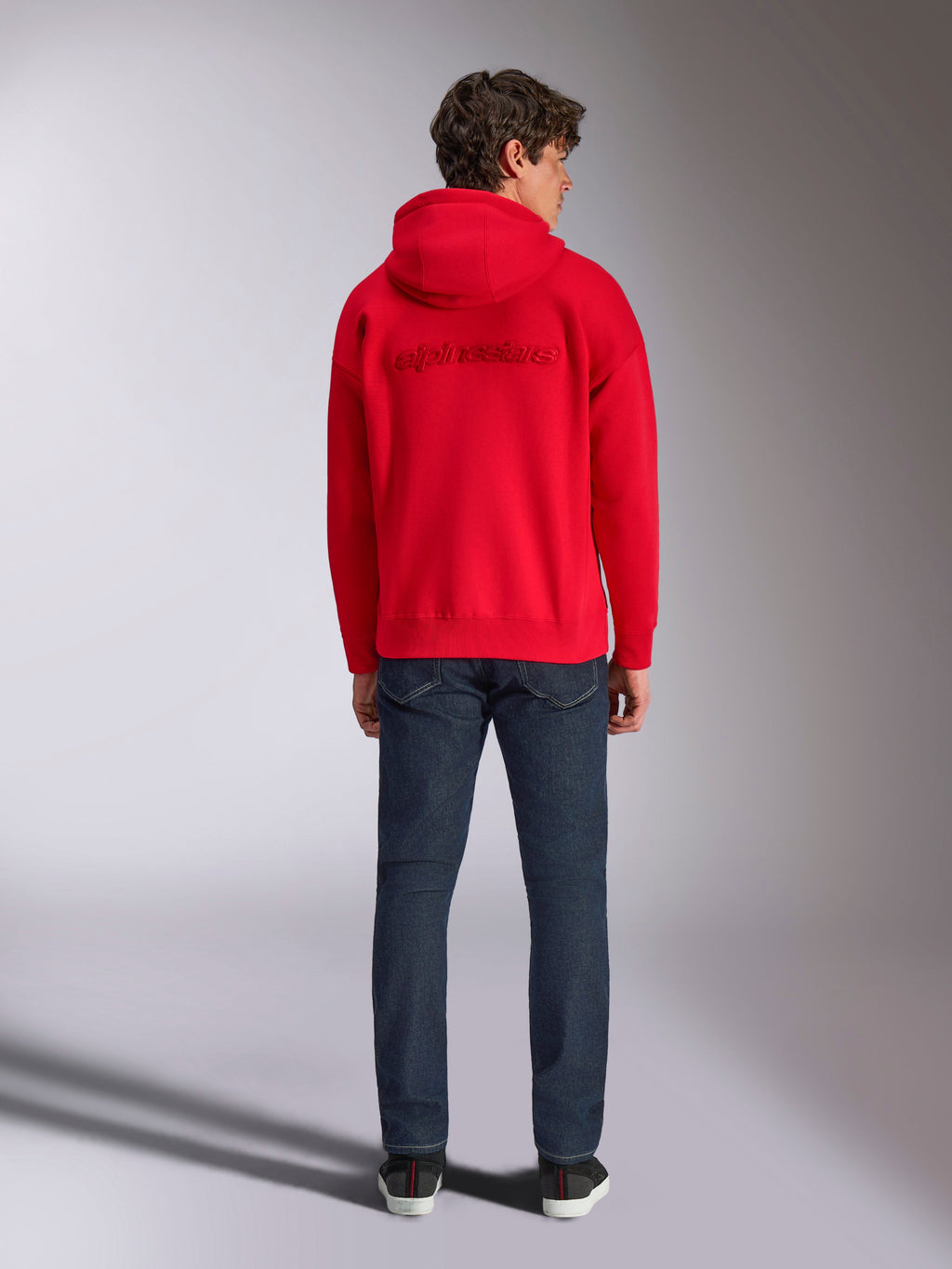 Alpinestars Emergent Oversized Hoodie, vermelho, vista traseira num modelo masculino, apresentando o logótipo Alpinestars bordado em 3D tom sobre tom, usado com calças de ganga azul escura, streetwear casual inspirado no desporto motorizado