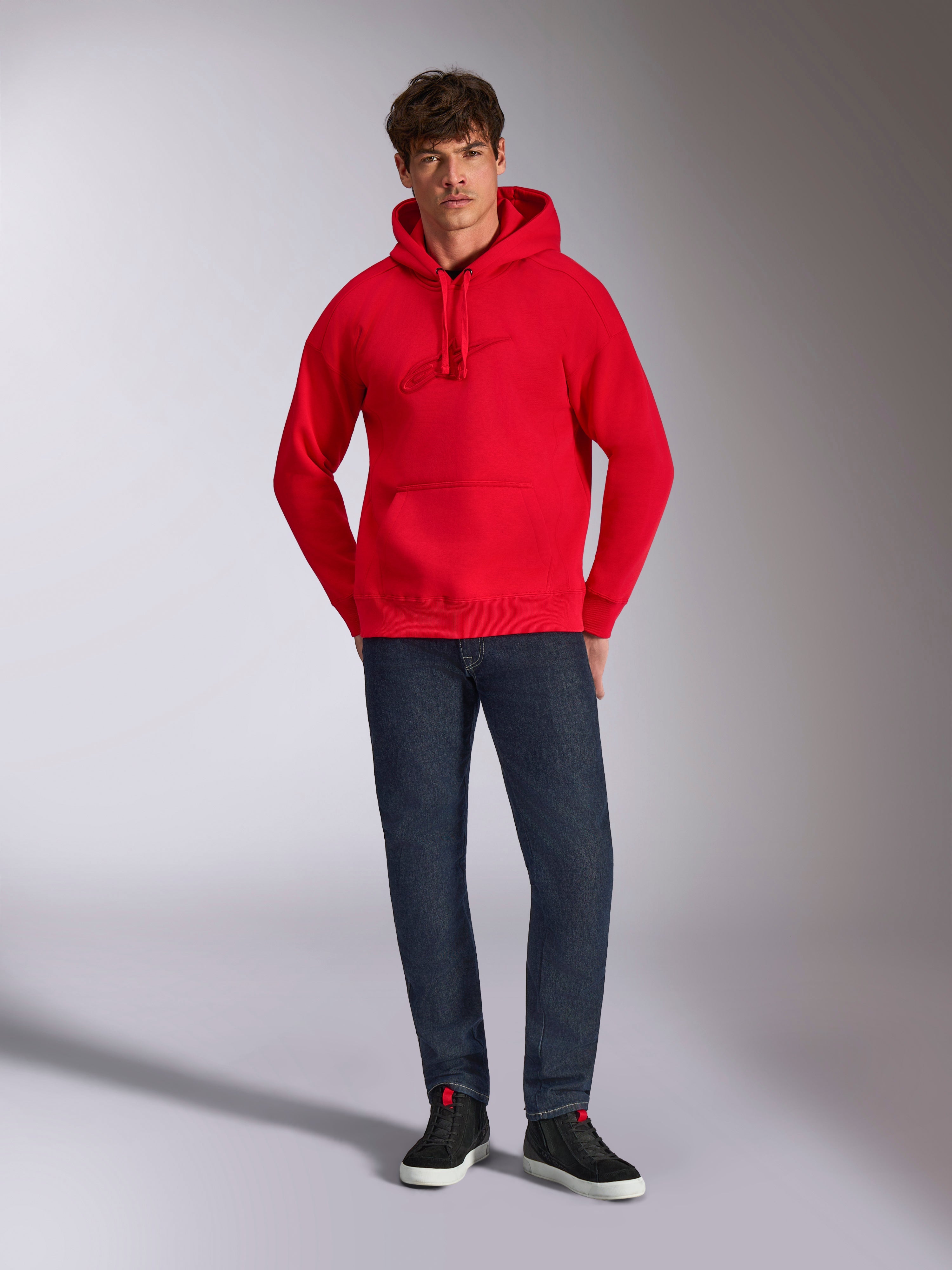 Alpinestars Emergent Oversized Hoodie, vermelho, com logótipo Astars bordado em 3D no peito no mesmo tom, bolso canguru e capuz com cordão, usado por um modelo com calças de ganga azul-escuro e sapatilhas pretas, estilo casual lifestyle streetwear