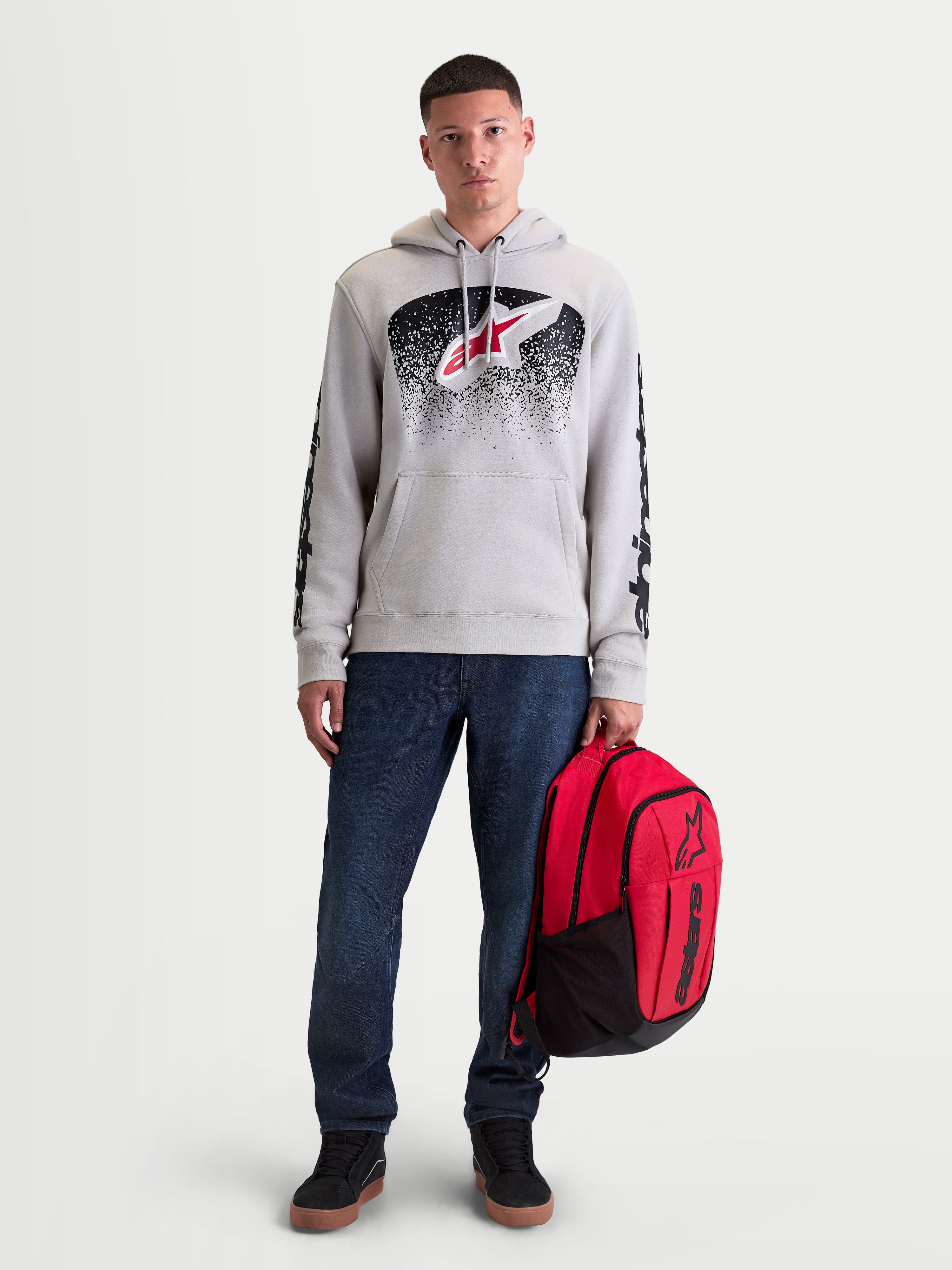 Alpinestars Camisola Com Capuz Obsessed, camisola de fleece para homem, cinzenta com gráficos inspirados em MX a vermelho e preto, logótipos nas mangas, modelo masculino a segurar mochila Alpinestars vermelha.