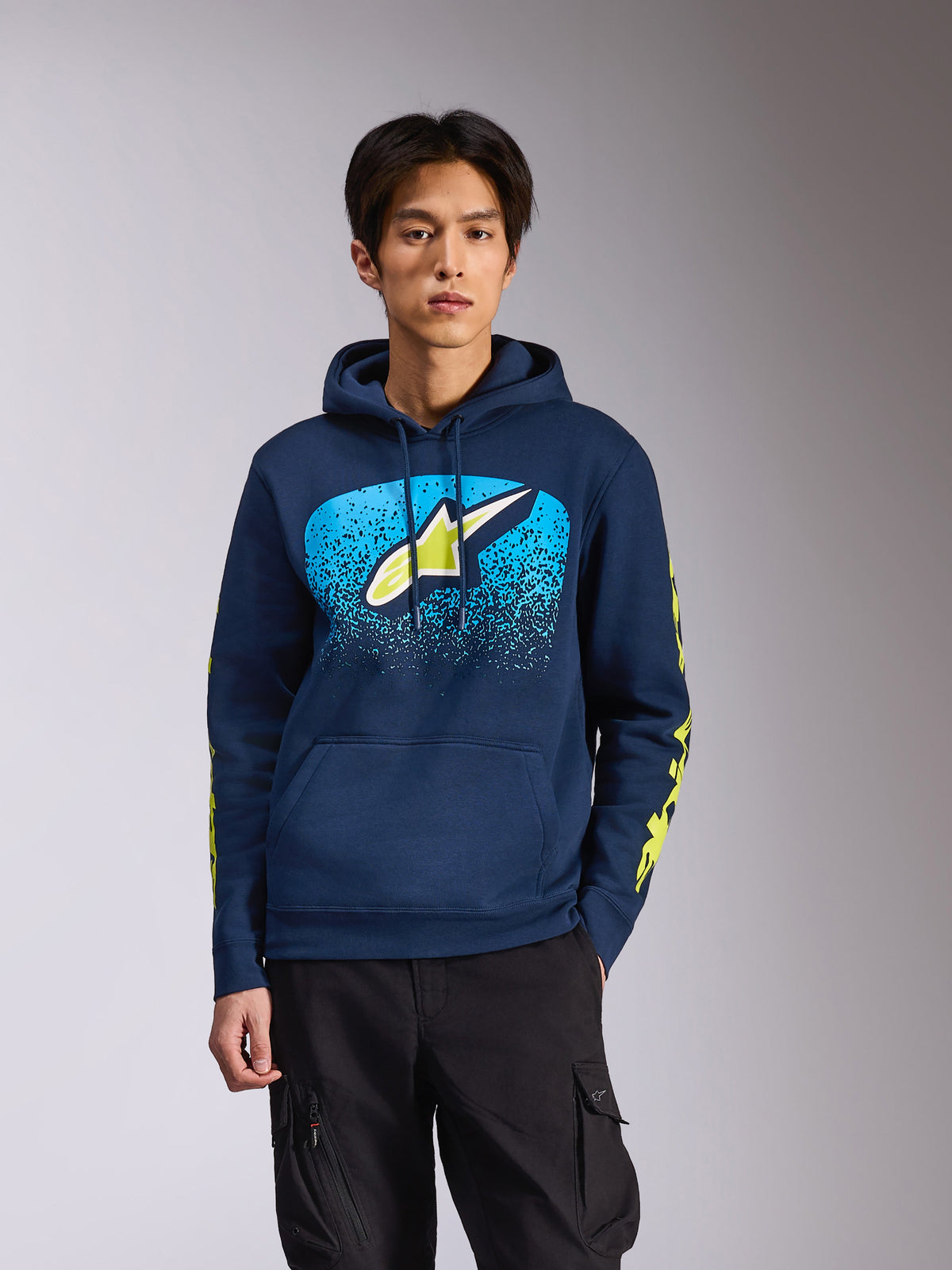 Alpinestars Obsessed Hoodie em azul-marinho, vestido por um modelo masculino, com um logótipo Astars verde-lima com gradação MX azul brilhante no peito e logótipos com o nome da marca em amarelo nas mangas, streetwear casual inspirado no desporto motorizado.