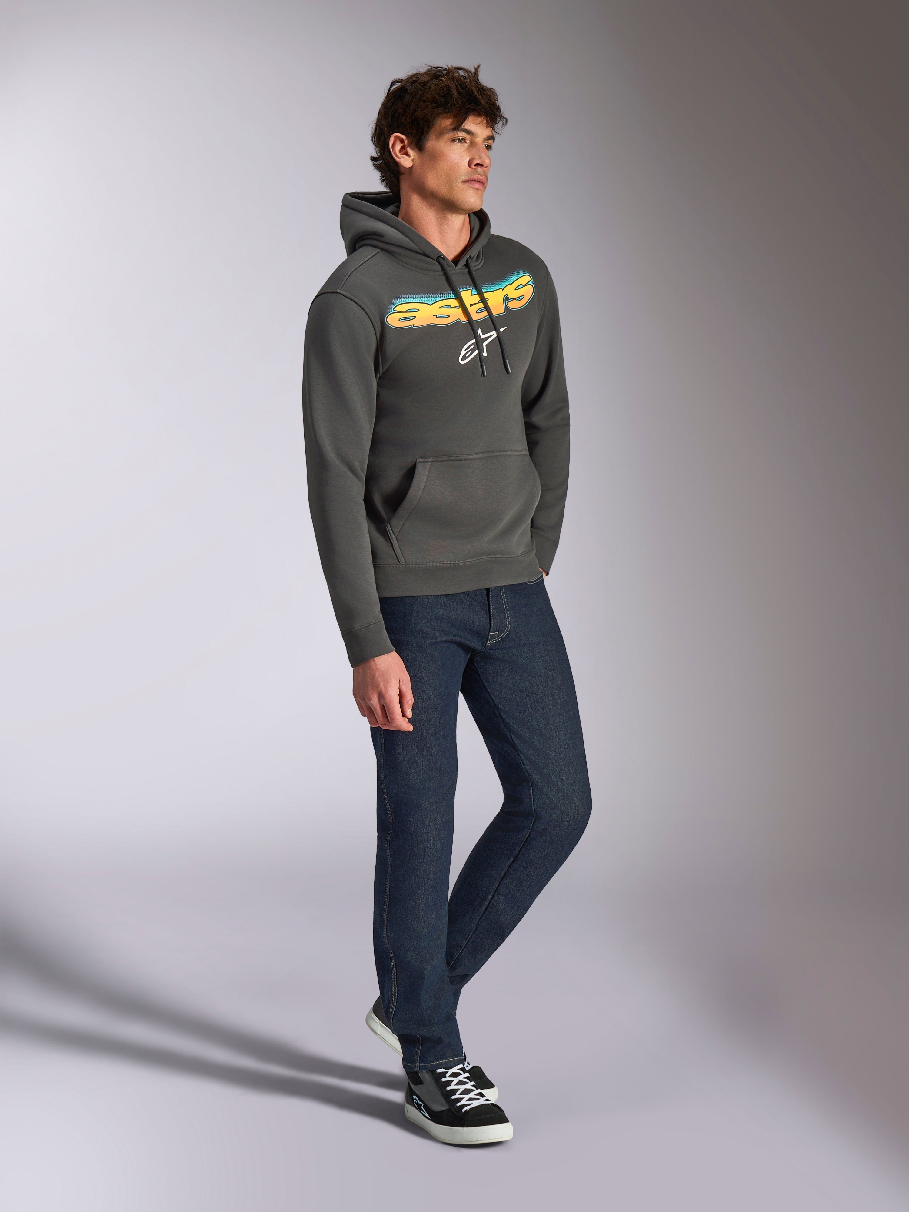 Alpinestars Ellipsoid Hoodie, Gunmetal gray pullover fleece, front curved Astars wordmark with blue and yellow color gradient and white logo, styled with dark jeans and black sneakers, casual streetwear.