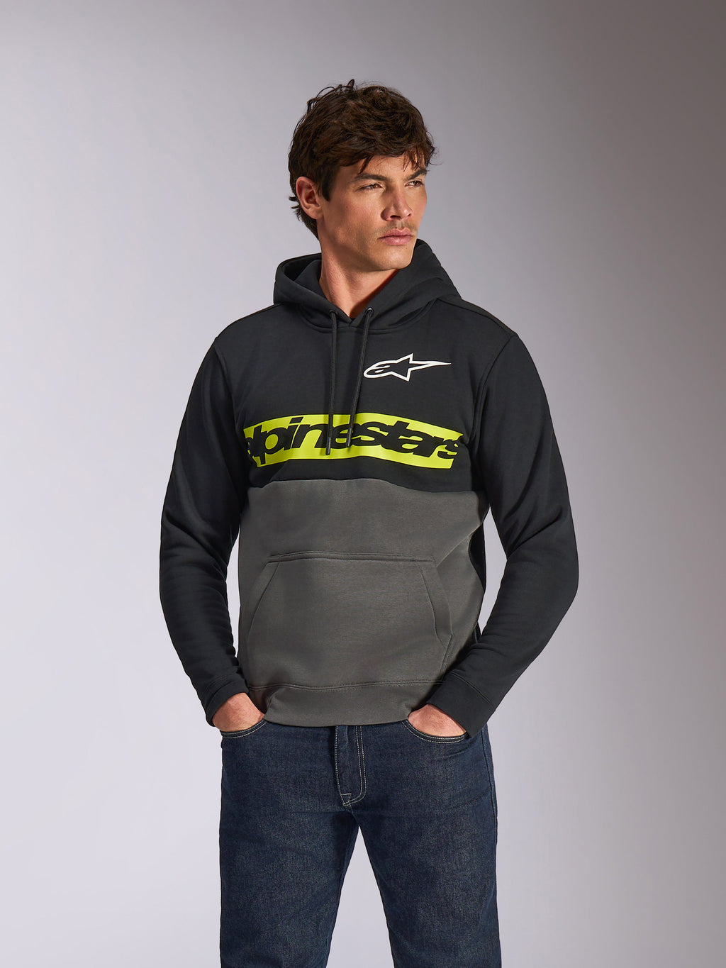 Alpinestars Parallel Hoodie, casual color-blocked pullover fleece, Black and Gunmetal with a vibrant yellow chest bar, Alpinestars word logo and white star logo, worn by a male model with dark blue jeans