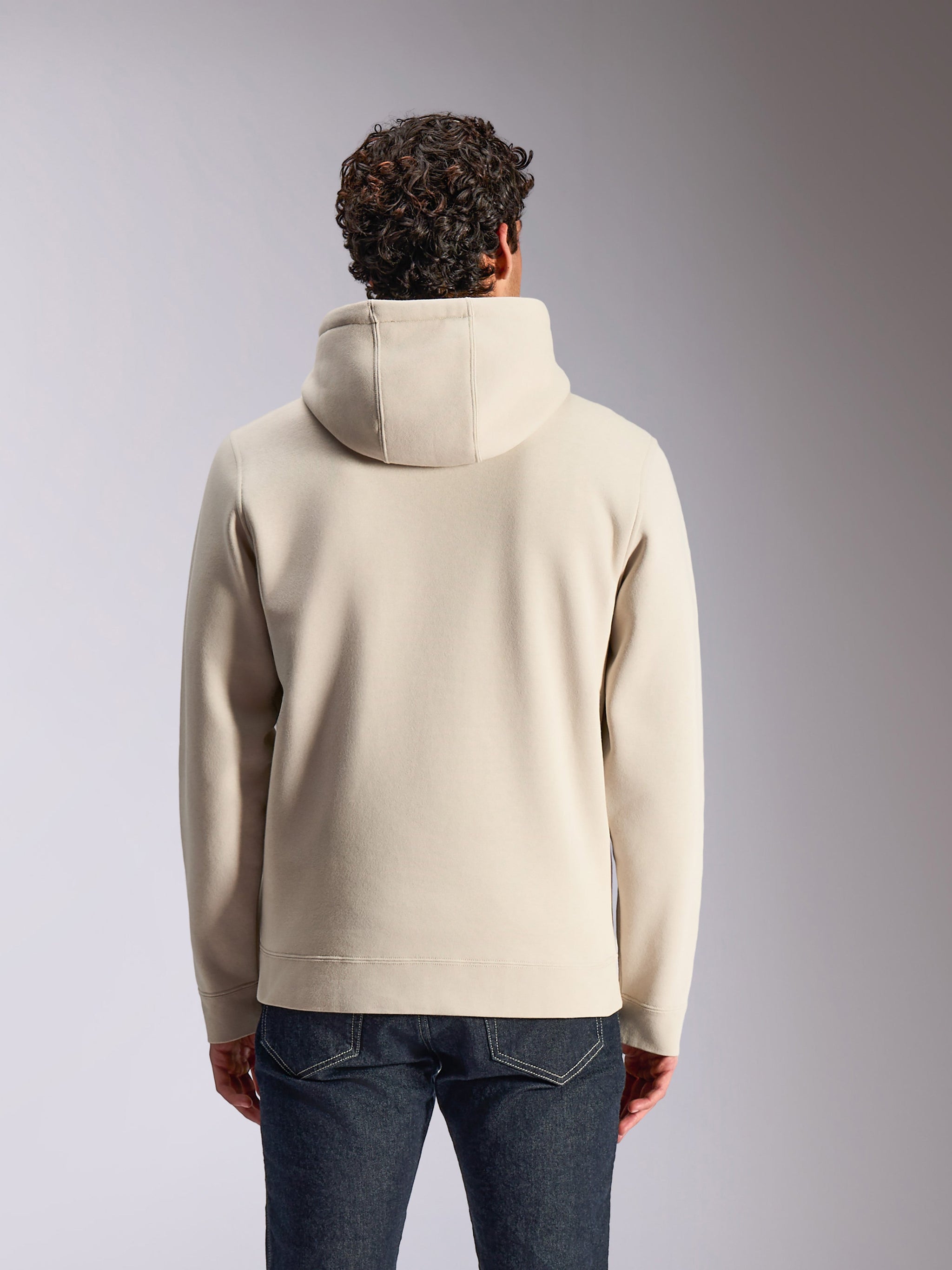 Alpinestars Pulse Hoodie, cor areia, vista traseira de um modelo masculino a usar a camisola lisa com capuz, combinada com calças de ganga escuras, streetwear casual inspirado no desporto motorizado