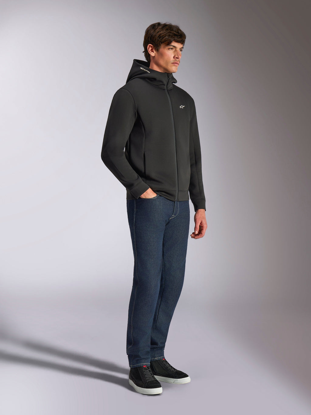 Alpinestars Cypher Hoodie, casual streetwear, black zip-up hoodie, male model wearing with dark blue jeans and black sneakers, high-neck hood with Alpinestars word logo, small white Astars logo on chest, sleek motorsport-inspired lifestyle look