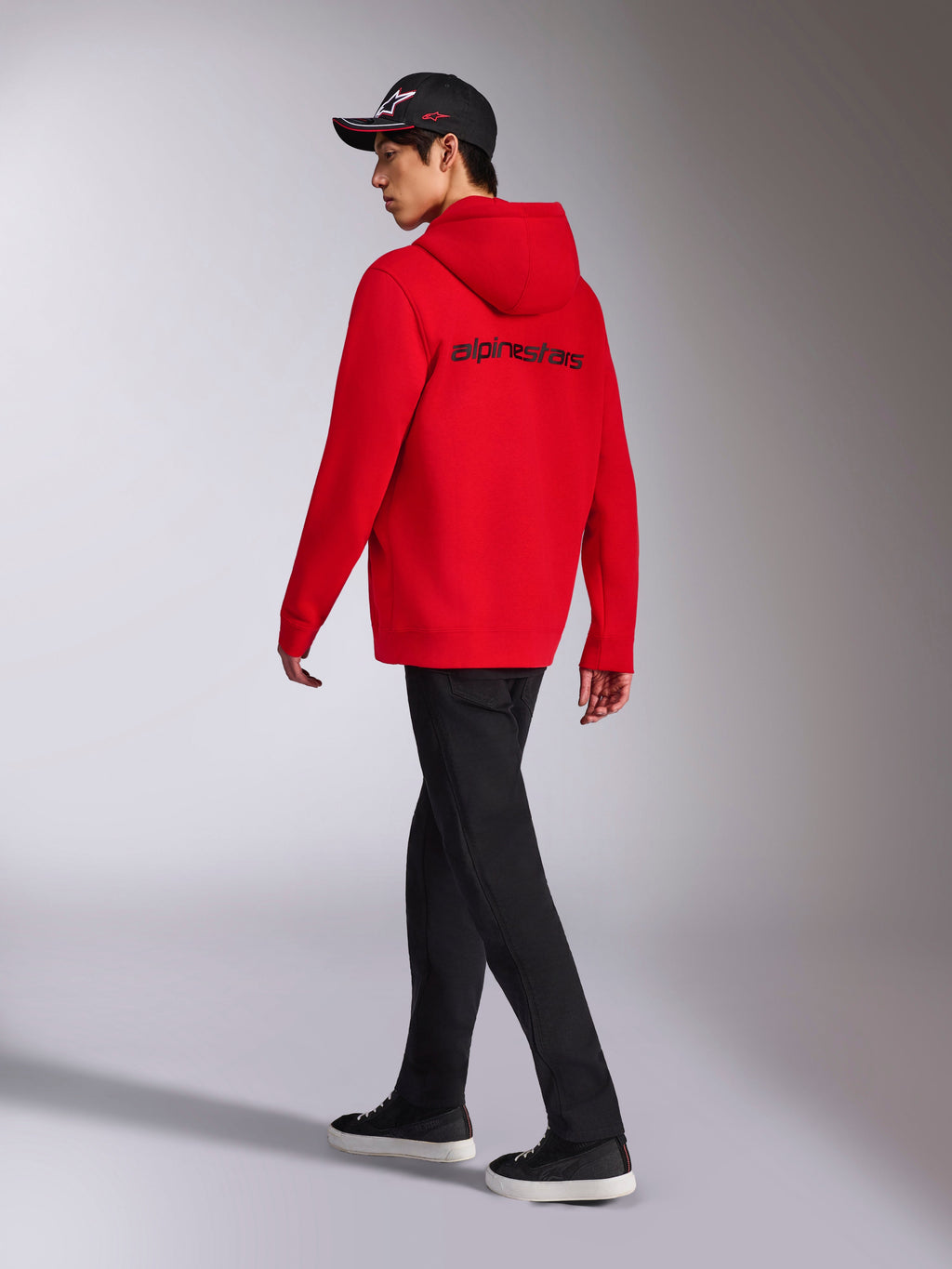Alpinestars Transverse Hoodie, sweatshirt vermelha com capuz e fecho de correr, vista traseira com logótipo Alpinestars a preto, usada por um modelo com calças pretas e boné preto com logótipo, streetwear casual inspirado no desporto motorizado