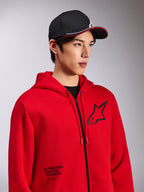 Alpinestars Transverse Hoodie, sweatshirt em malha polar vermelha com fecho frontal, logótipo Astars preto no peito e texto 'One Goal. One Vision.' na parte inferior frontal, streetwear casual de motorsport, usada por um modelo com um boné preto