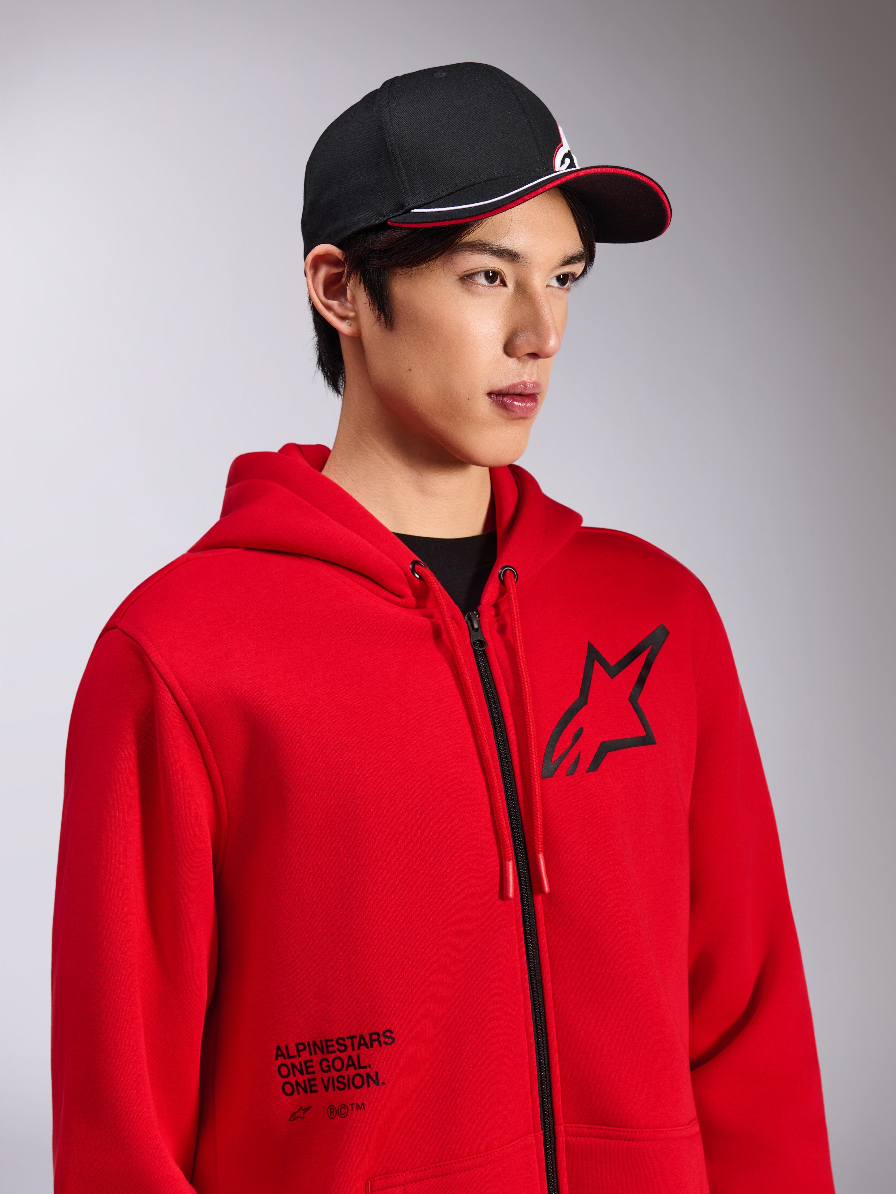 Alpinestars Transverse Hoodie, sweatshirt em malha polar vermelha com fecho frontal, logótipo Astars preto no peito e texto 'One Goal. One Vision.' na parte inferior frontal, streetwear casual de motorsport, usada por um modelo com um boné preto