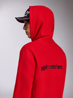 Alpinestars Transverse Hoodie, sweatshirt com capuz em vermelho, vista lateral traseira mostrando o logótipo Alpinestars a preto nas costas, usada com o capuz colocado e um boné preto, streetwear casual inspirado no desporto motorizado