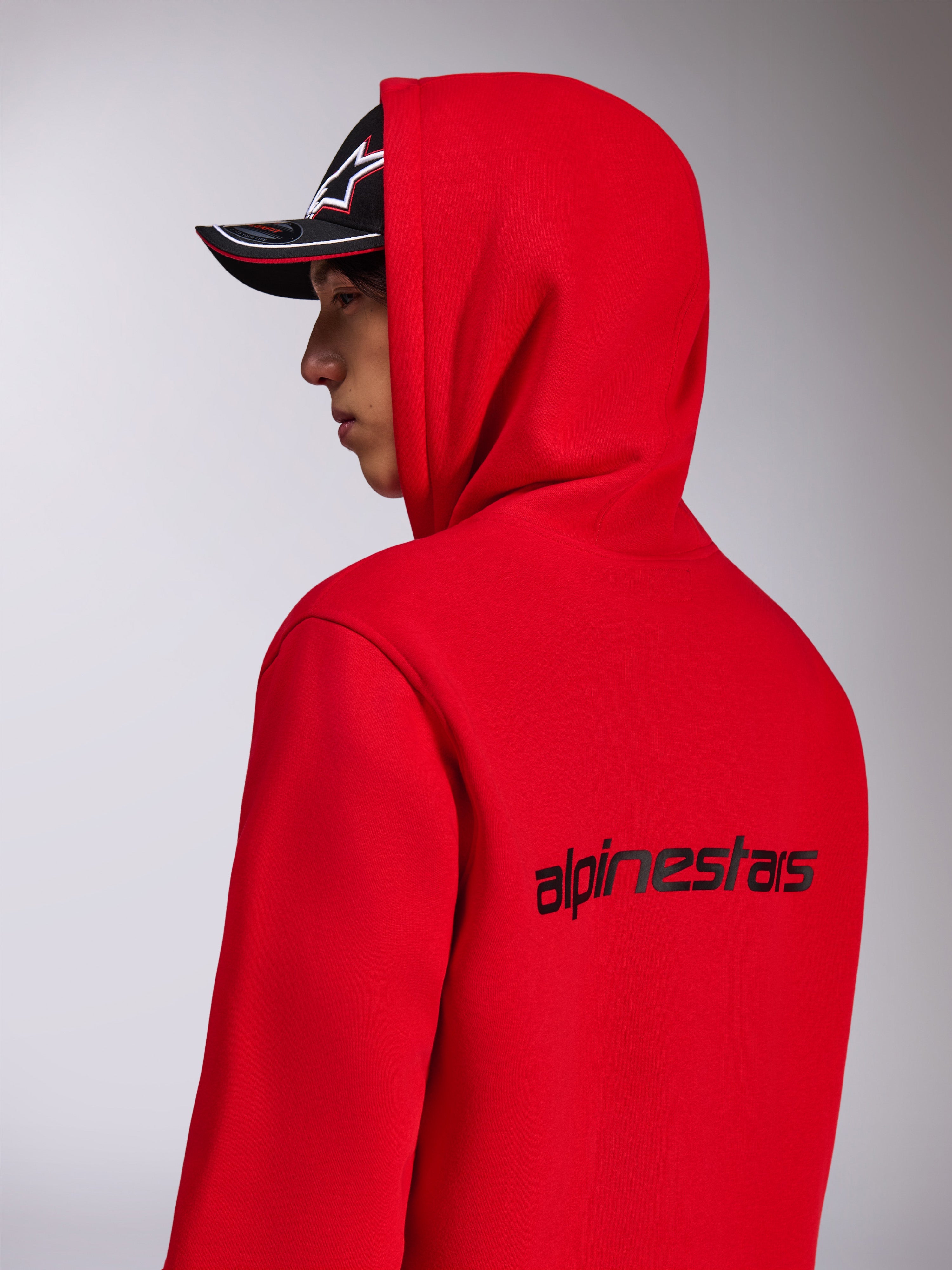 Alpinestars Transverse Hoodie, sweatshirt com capuz em vermelho, vista lateral traseira mostrando o logótipo Alpinestars a preto nas costas, usada com o capuz colocado e um boné preto, streetwear casual inspirado no desporto motorizado