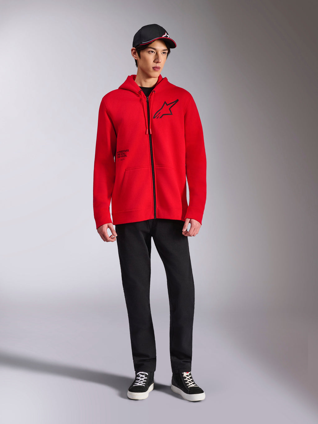 Alpinestars Transverse Hoodie, camisola com capuz em tecido polar vermelho com fecho de correr, logótipo Astars preto no peito e impressão One Goal na lateral, usada por modelo masculino com calças pretas e boné, streetwear casual inspirado no desporto motorizado