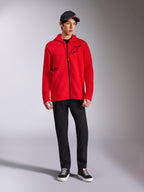 Alpinestars Transverse Hoodie, camisola com capuz em tecido polar vermelho com fecho de correr, logótipo Astars preto no peito e impressão One Goal na lateral, usada por modelo masculino com calças pretas e boné, streetwear casual inspirado no desporto motorizado
