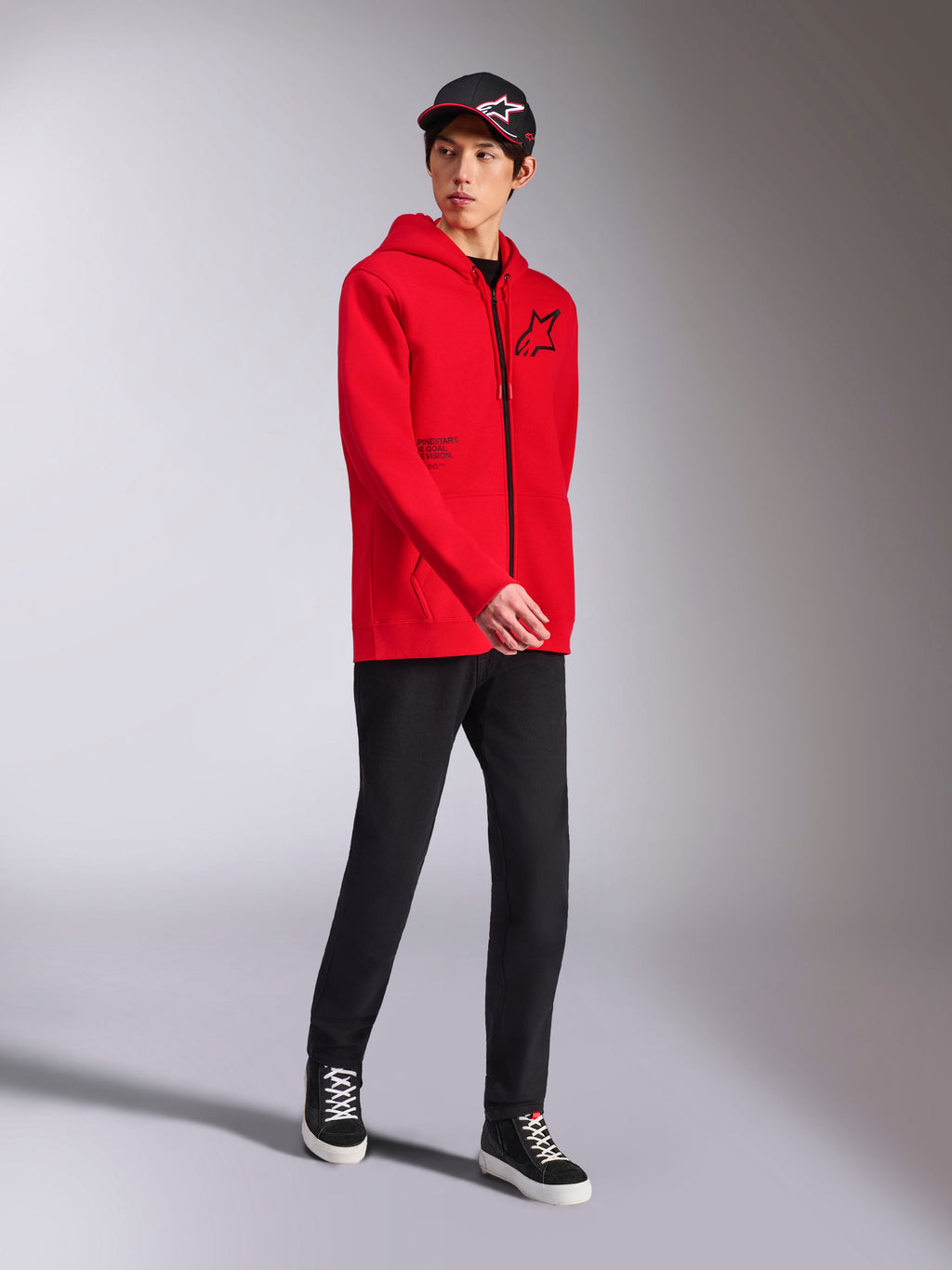 Alpinestars Transverse Hoodie em vermelho, modelo masculino a usar sweatshirt com capuz e fecho integral em tecido polar, logótipo Astars e texto impressos a preto, combinado com calças e boné pretos, estilo casual motorsport streetwear