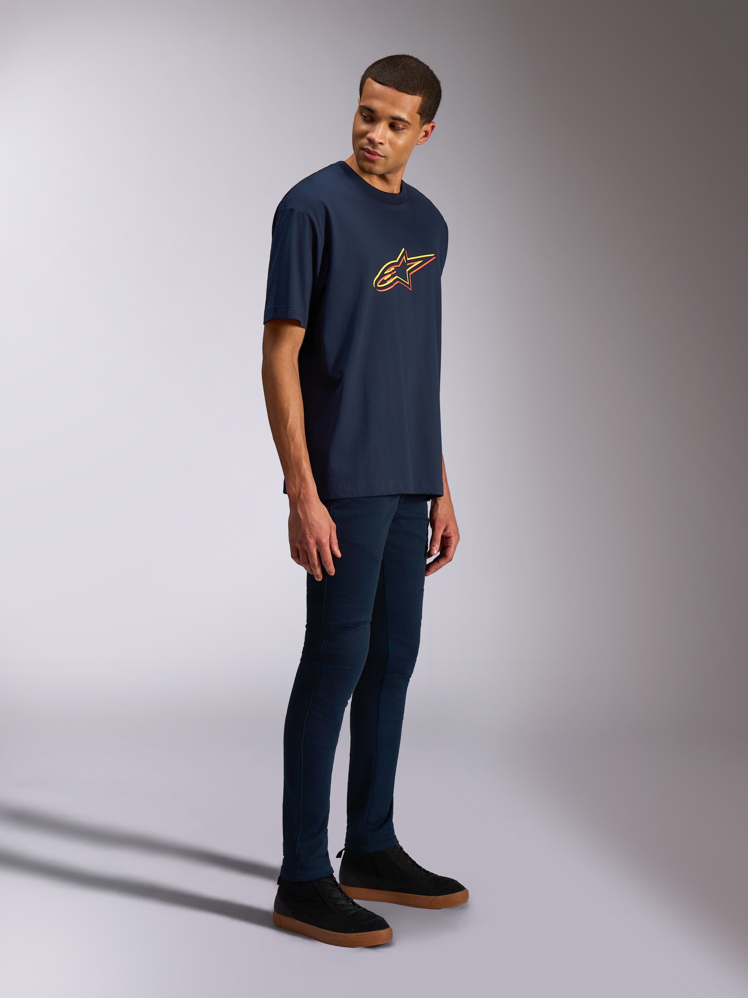 Alpinestars Alter Oversized CSF Tee, t-shirt de manga curta, azul-marinho, logótipo Astars amarelo e laranja, usada por um modelo com calças de ganga escuras e sapatilhas pretas, streetwear casual inspirado no desporto motorizado