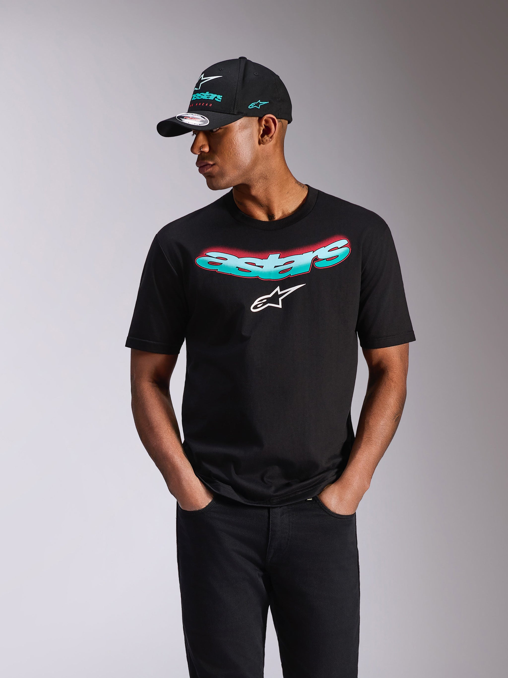 Alpinestars Ellipsoid CSF Tee, T-shirt de manga curta, preta, palavra Astars com gradiente azul-turquesa e rosa e logótipo branco, usada por um modelo com boné Alpinestars a condizer e calças de ganga pretas, streetwear casual de desportos motorizados