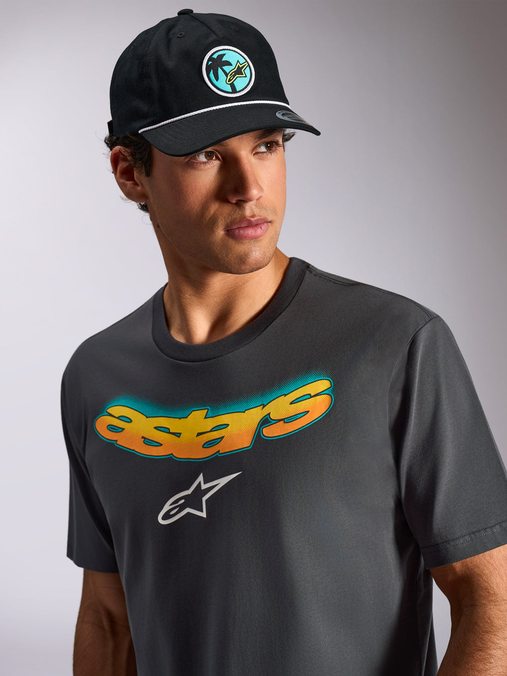 T-shirt Alpinestars Ellipsoid CSF de manga curta, cinzento escuro, com a palavra "Astars" curvada em gradiente amarelo e laranja e o logótipo Astars branco, vestida por um modelo com boné preto, estilo de vida casual motorsport streetwear