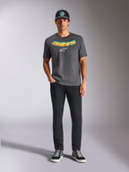 T-shirt Alpinestars Ellipsoid CSF, t-shirt de manga curta, cinzento carvão, com uma inscrição curva em degradê amarelo e cor de laranja e logótipo Astars branco, usada por um modelo com calças pretas e boné, estilo sportswear casual