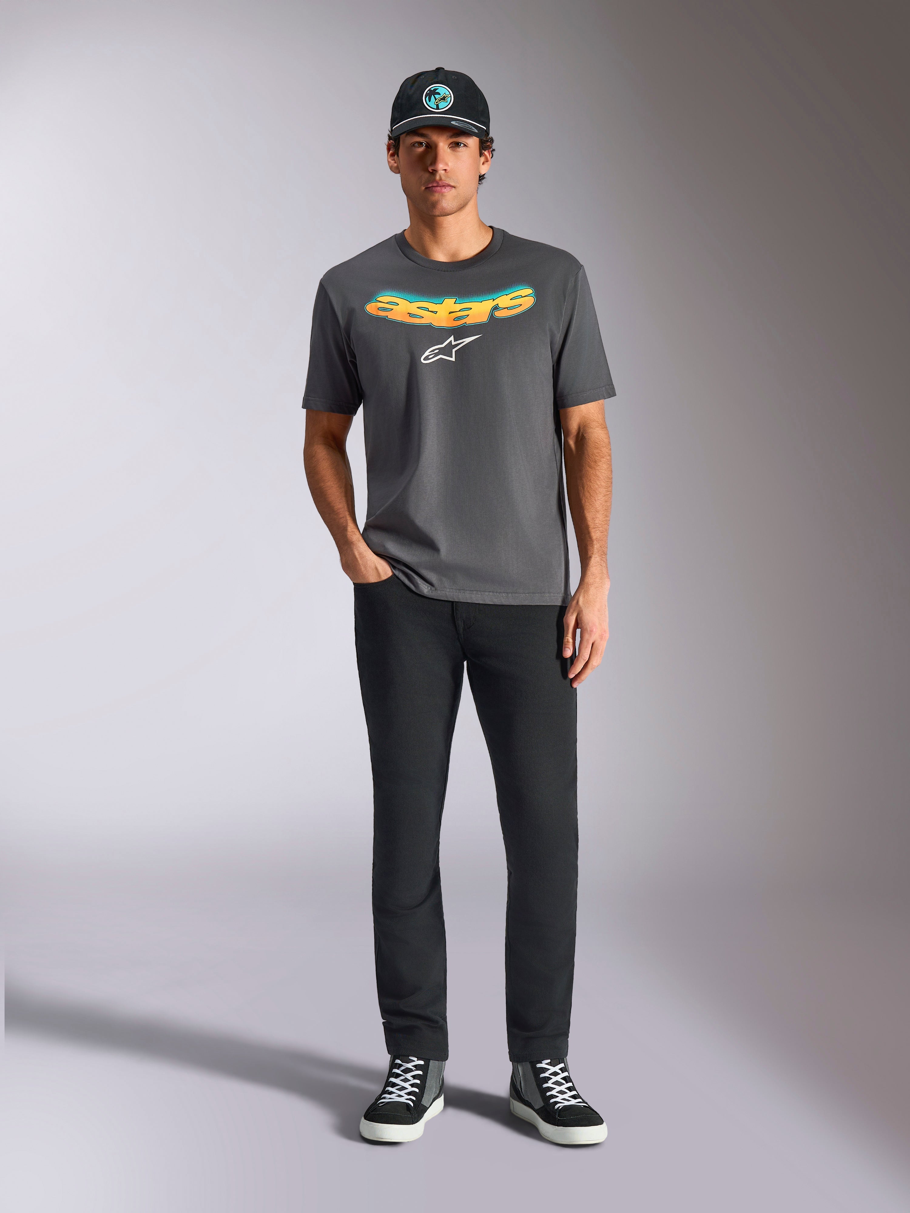 T-shirt Alpinestars Ellipsoid CSF, t-shirt de manga curta, cinzento carvão, com uma inscrição curva em degradê amarelo e cor de laranja e logótipo Astars branco, usada por um modelo com calças pretas e boné, estilo sportswear casual
