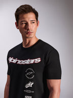 T-shirt Alpinestars Abus CSF, t-shirt de manga curta preta usada por um modelo, com logótipo Alpinestars curvo em branco com contorno vermelho e vários logótipos gráficos sobrepostos no peito, estilo streetwear casual de desporto motorizado