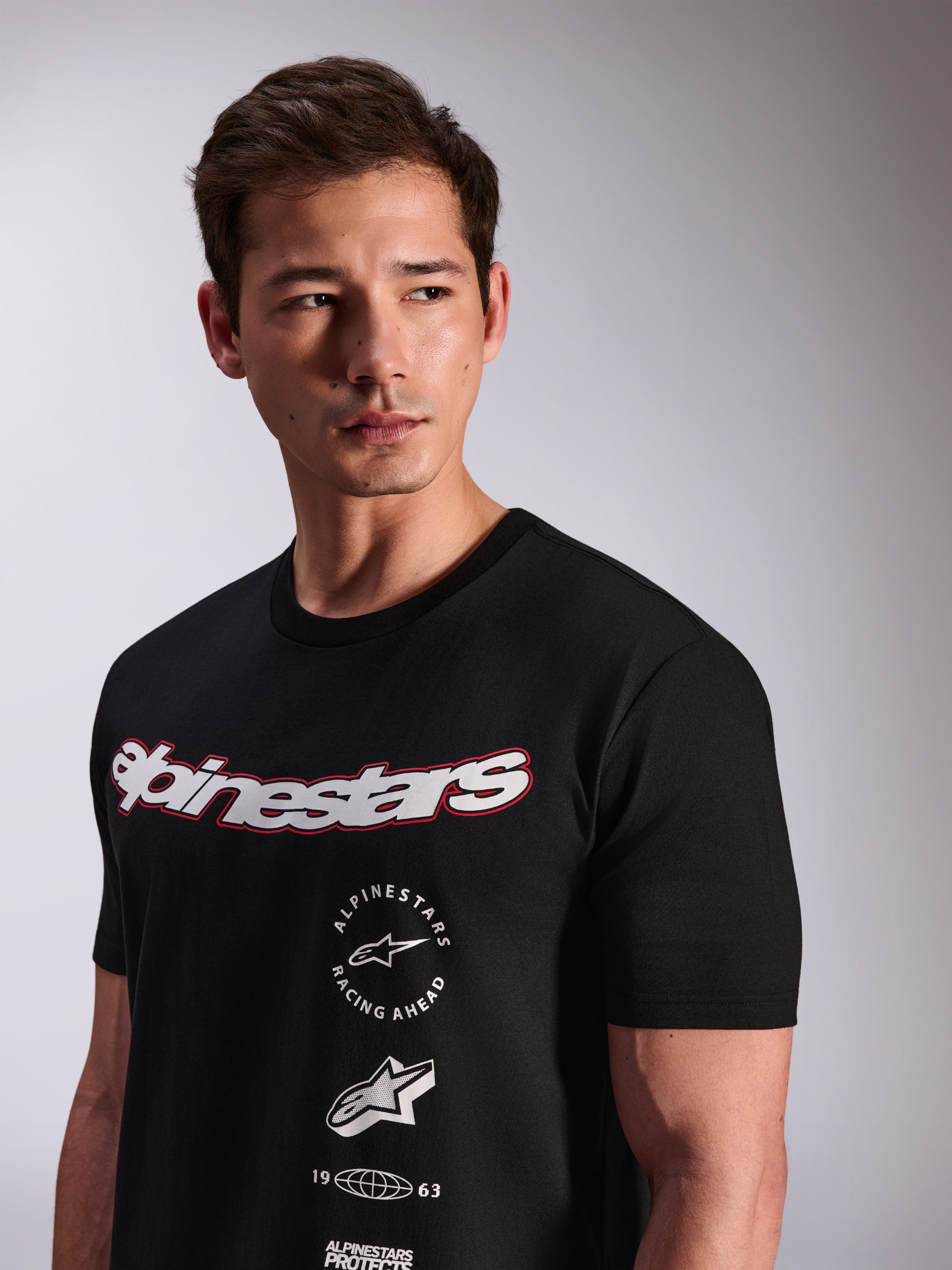 T-shirt Alpinestars Abus CSF, t-shirt de manga curta preta usada por um modelo, com logótipo Alpinestars curvo em branco com contorno vermelho e vários logótipos gráficos sobrepostos no peito, estilo streetwear casual de desporto motorizado