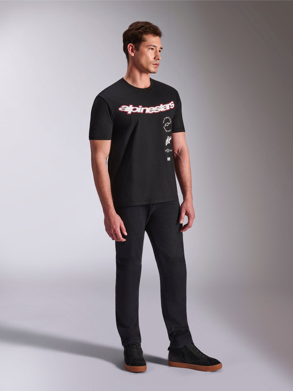 Alpinestars Abus CSF Tee, T-shirt de manga curta, preta, com logótipo Alpinestars curvo em vermelho e branco e gráficos empilhados verticalmente, usada com calças pretas e sapatilhas, streetwear casual inspirado no desporto motorizado