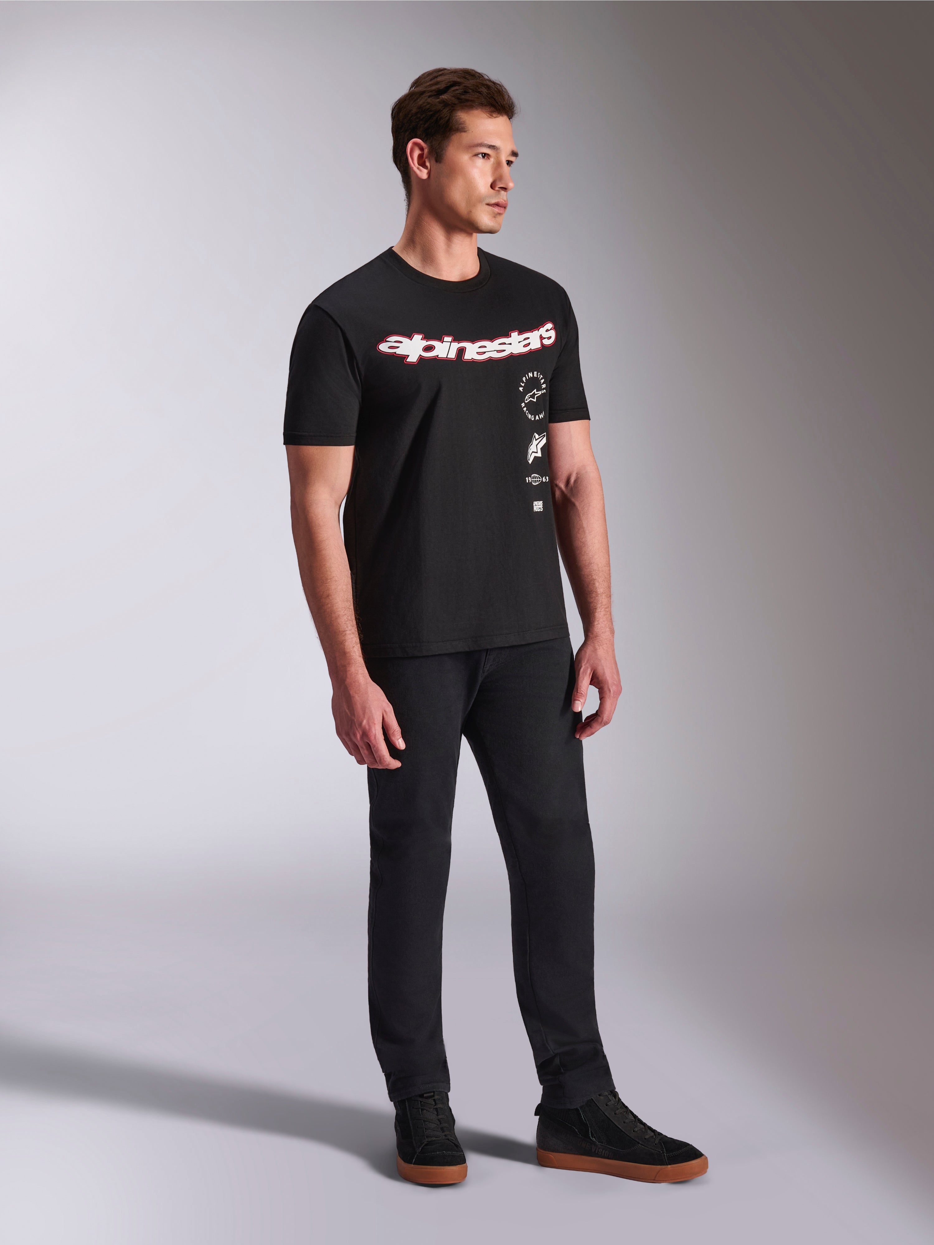 Alpinestars Abus CSF Tee, T-shirt de manga curta, preta, com logótipo Alpinestars curvo em vermelho e branco e gráficos empilhados verticalmente, usada com calças pretas e sapatilhas, streetwear casual inspirado no desporto motorizado
