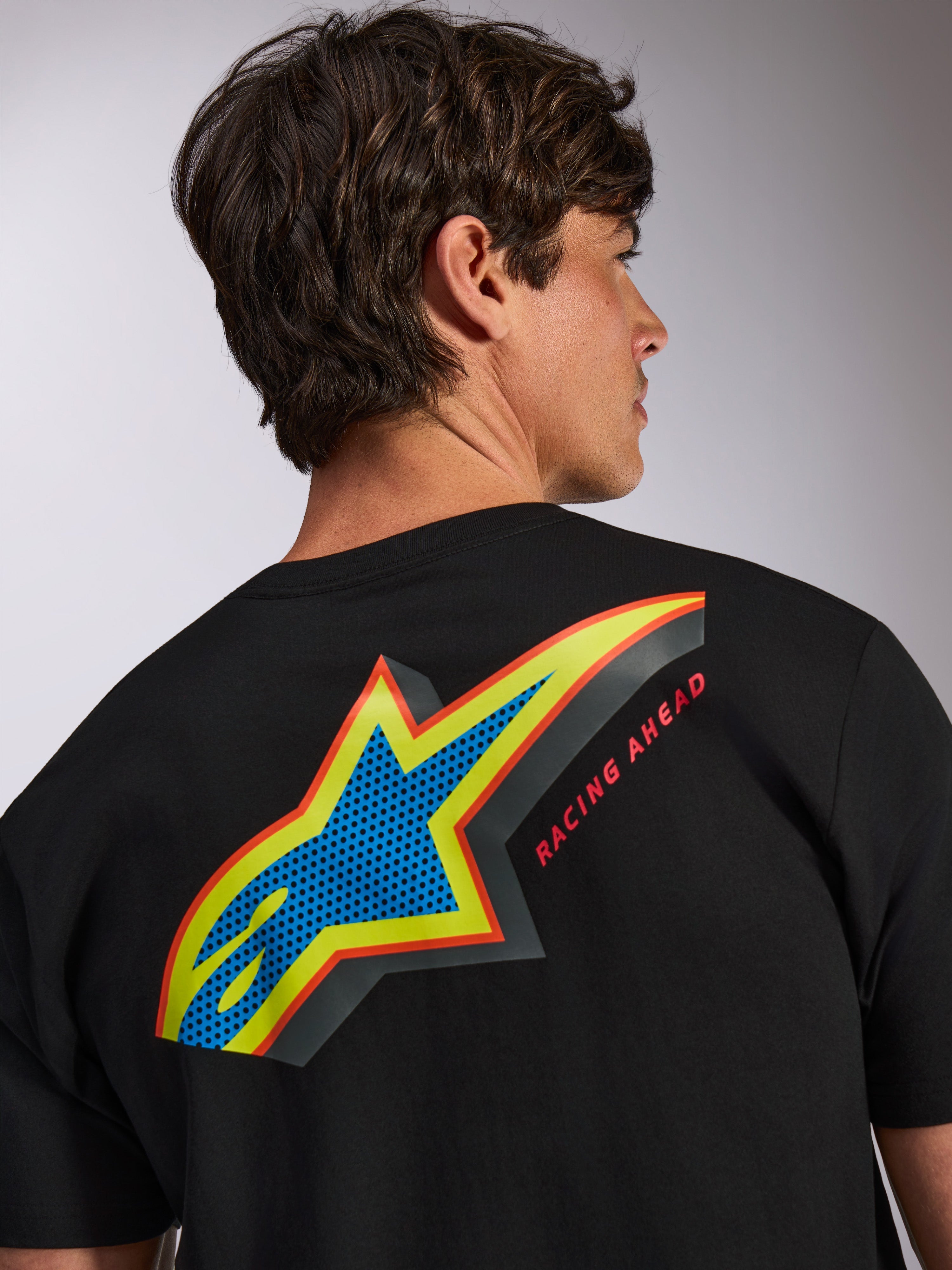 Alpinestars Angler CSF Tee, t-shirt de manga curta preta, logótipo Astars grande e multicolor nas costas com preenchimento perfurado azul, contornos amarelos e vermelhos e texto 'RACING AHEAD', vestuário casual de inspiração motorsport