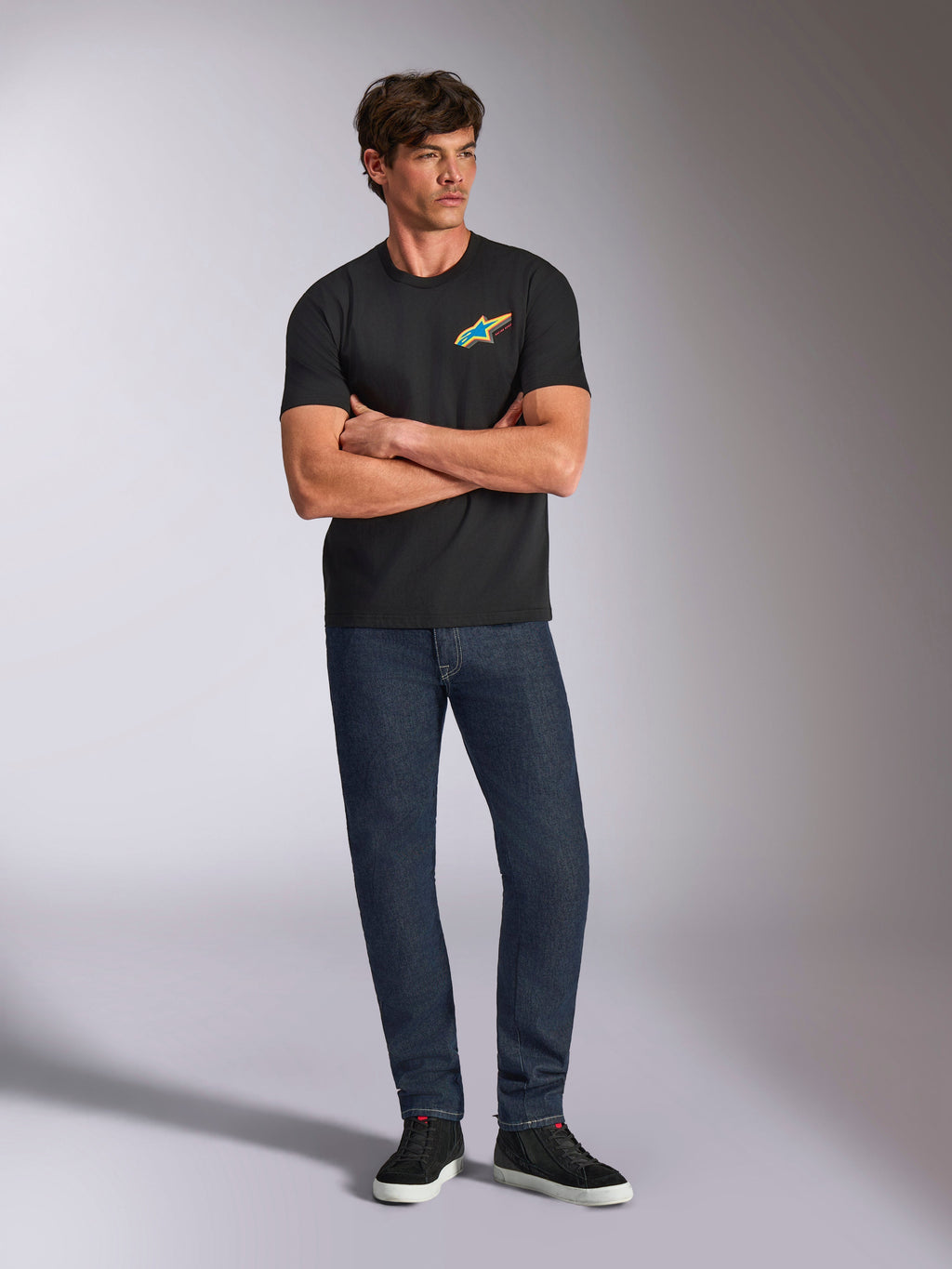 Alpinestars Angler CSF Tee, t-shirt de manga curta, preta, logótipo Astars vibrante e multicolor no peito, usada por modelo com os braços cruzados, calças de ganga escura e ténis pretos, streetwear casual de motorsport