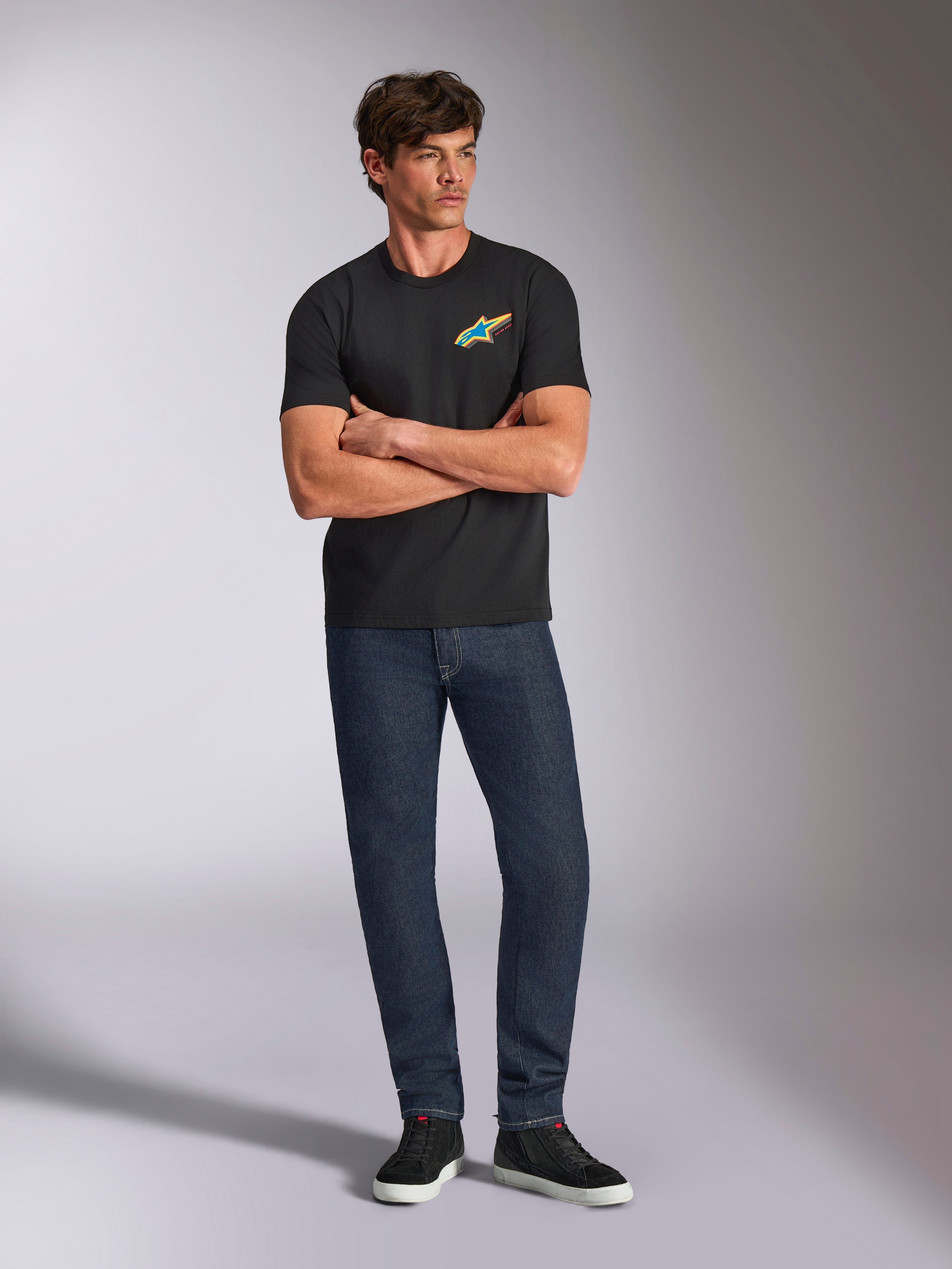 Alpinestars Angler CSF Tee, t-shirt de manga curta, preta, logótipo Astars vibrante e multicolor no peito, usada por modelo com os braços cruzados, calças de ganga escura e ténis pretos, streetwear casual de motorsport