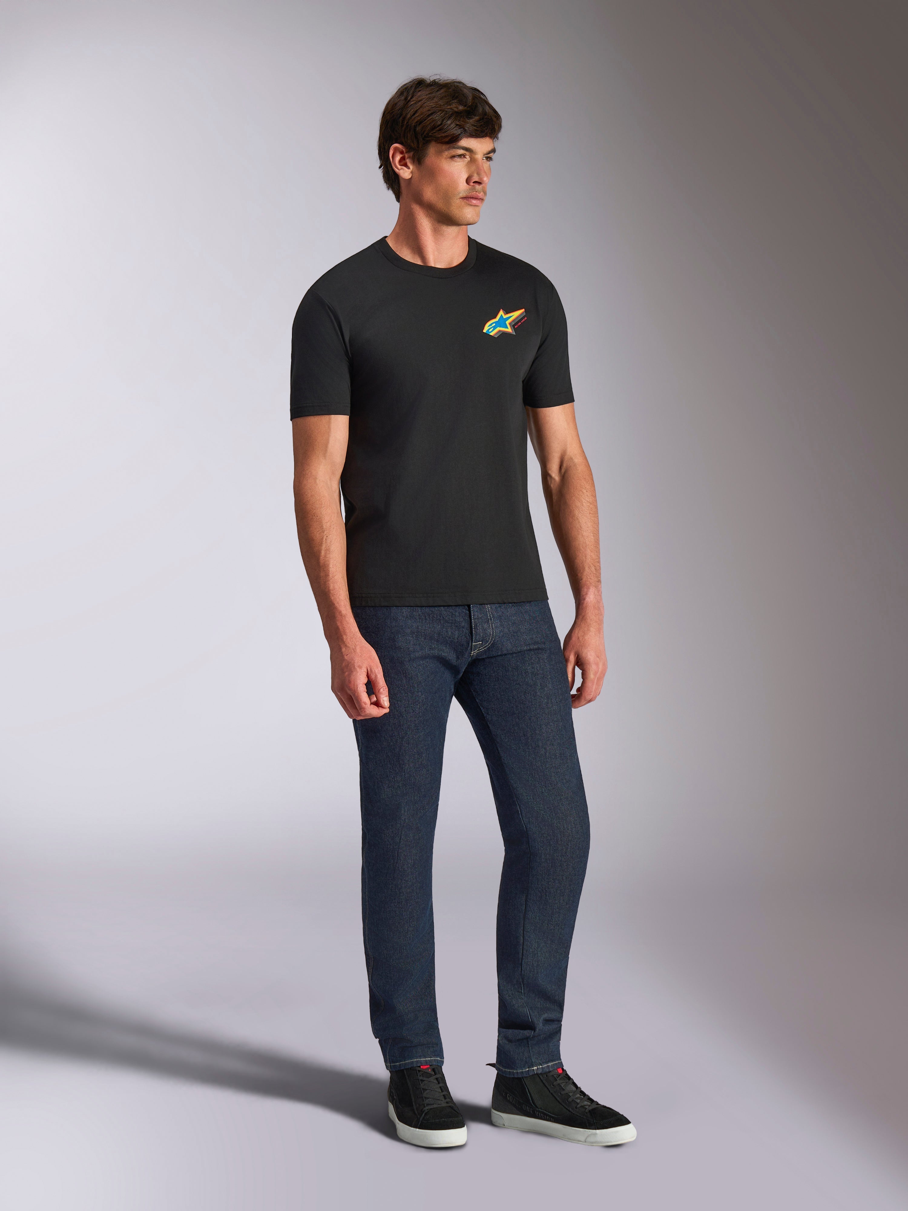 Alpinestars Angler CSF Tee, t-shirt de manga curta preta com logótipo Astars vibrante e multicolor no peito, modelo masculino com calças de ganga azul escura e sapatilhas pretas, vestuário casual de estilo streetwear e motorsport