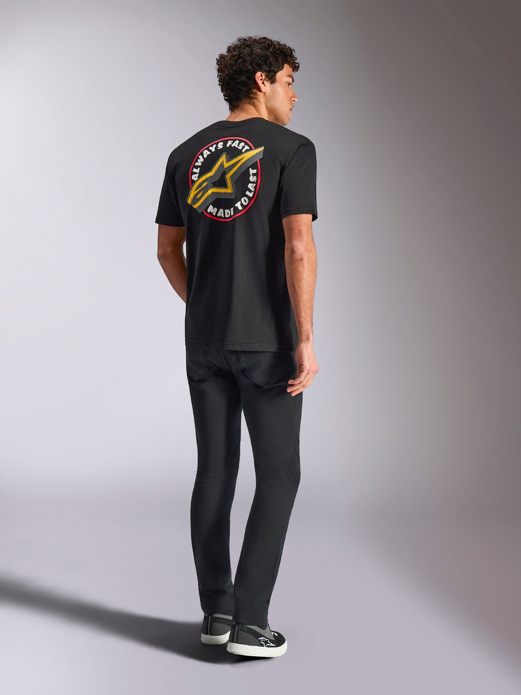Alpinestars Aorbit CSF Tee, T-shirt de manga curta, preta, vista traseira com um logótipo Astars grande e amarelo e o gráfico circular 'Always Fast Made to Last', usada com calças de ganga pretas e sapatilhas casuais, estilo streetwear inspirado no desporto motorizado