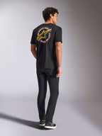 Alpinestars Aorbit CSF Tee, T-shirt de manga curta, preta, vista traseira com um logótipo Astars grande e amarelo e o gráfico circular 'Always Fast Made to Last', usada com calças de ganga pretas e sapatilhas casuais, estilo streetwear inspirado no desporto motorizado